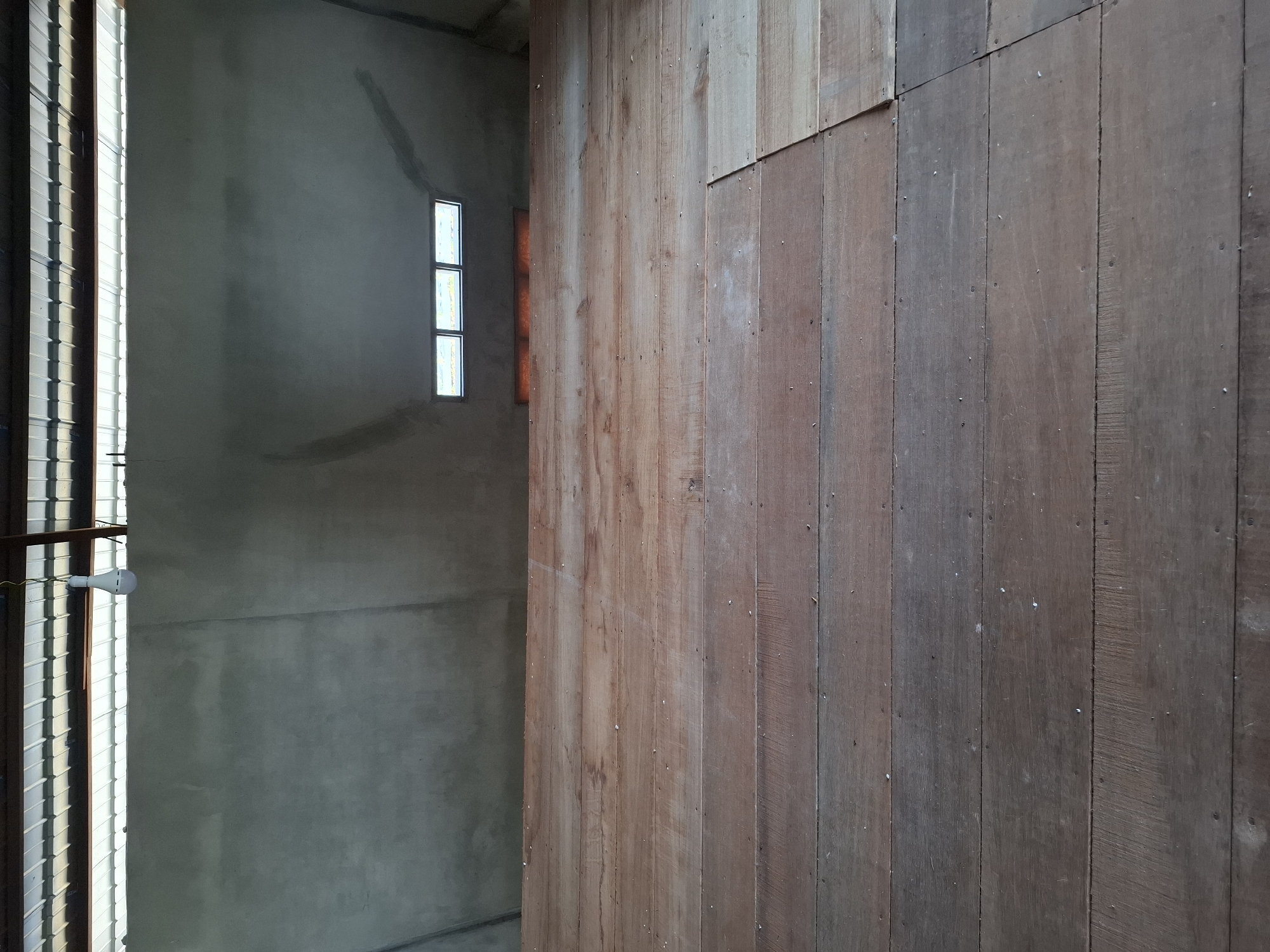 The image shows the interior of a building under construction, likely in Gronggrong, Indonesia. The foreground is dominated by a wooden floor made of planks, some of which appear to be unfinished or rough. The floor has a rustic appearance with visible nail heads and wood grain. In the background, there is a concrete wall that extends upwards to a corrugated metal roof. A single light bulb hangs from the ceiling. Towards the top of the concrete wall, three small rectangular windows are visible, allowing some natural light to enter. The overall scene suggests a functional, possibly agricultural or storage space, in an early stage of development.