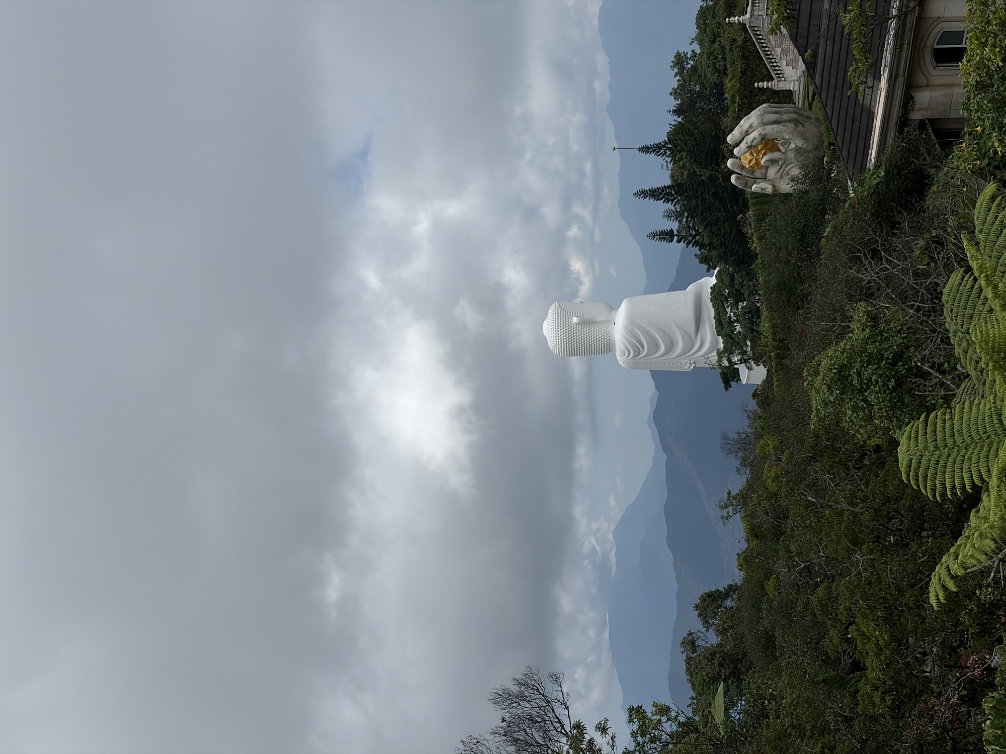 The image shows a partial view of the Ba Na Hills complex in Đà Nẵng, Vietnam. The primary focus is on a large, white Buddha statue standing against a backdrop of distant mountains partially obscured by clouds. To the right of the Buddha, a sculpted pair of stone hands hold a golden object, located near a building with ornate details. Lush green foliage and trees cover the hillside, framing the statue and architecture. The sky is overcast with heavy clouds.