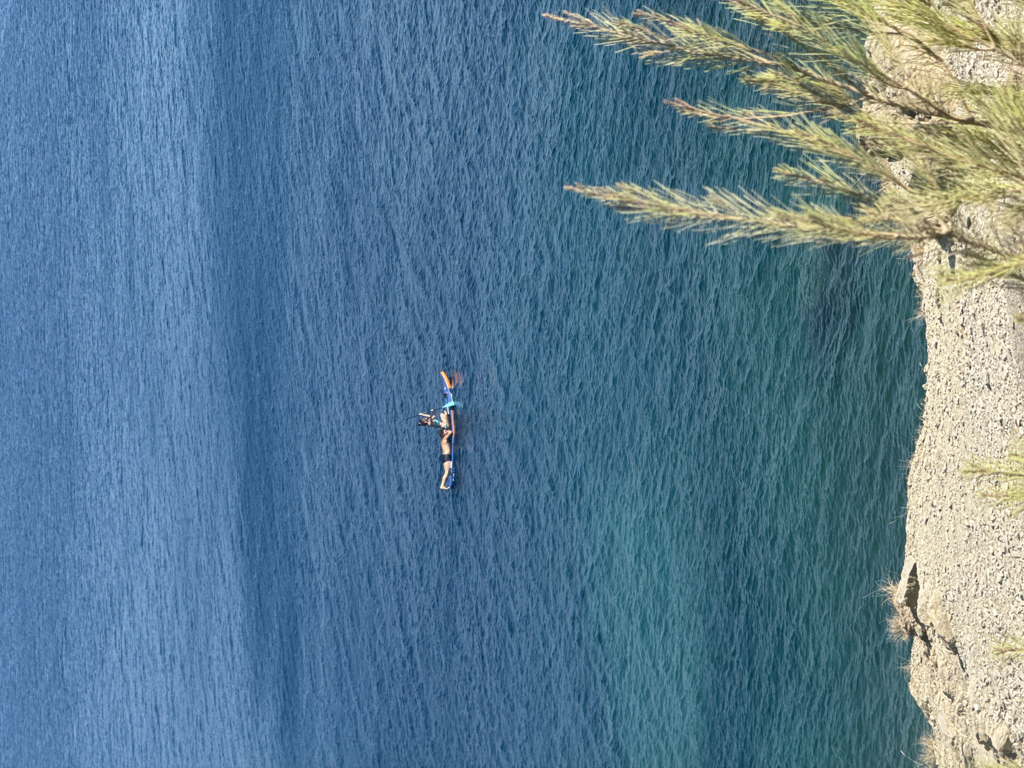In the city of Sutomore, Montenegro, a person is lying prone on a stand-up paddleboard in the Adriatic Sea. The water is a deep blue, transitioning to a lighter turquoise near the shore. The paddleboard is orange and blue. Another person is partially visible behind the first, seated on the paddleboard and holding a paddle. The foreground shows a rocky, sparsely vegetated shoreline.