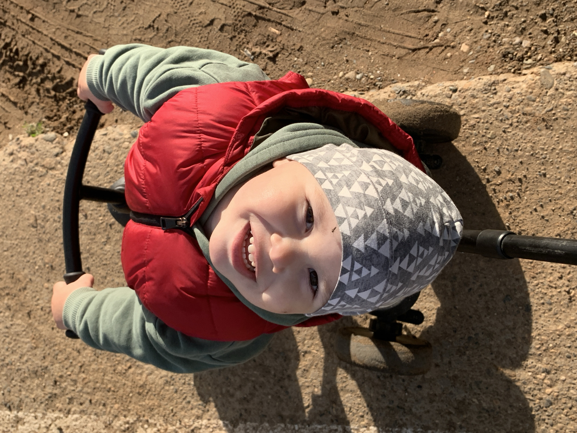 A high-angle, top-down shot captures a young child, likely a toddler, smiling broadly while seated on a ride-on toy. The child looks directly into the camera with a wide, open-mouthed grin, revealing their teeth. They are dressed in a light grey beanie with a white geometric triangle pattern, a light green hooded sweatshirt, and a bright red puffer vest. Their hands are visible gripping black handlebars, which are part of the toy.
The scene is set outdoors on a sunny day. The ground beneath and around the child is a mix of dirt and small gravel, with distinct tire tracks visible in the dirt, suggesting a path or play area. A small portion of a dark wheel from the toy is visible on the right. Long shadows are cast on the ground, indicating strong, direct sunlight, likely during mid-morning or afternoon. The child's joyful expression conveys happiness and excitement. No visible text is present in the image. The location is contextualized as Фоки, Russia.