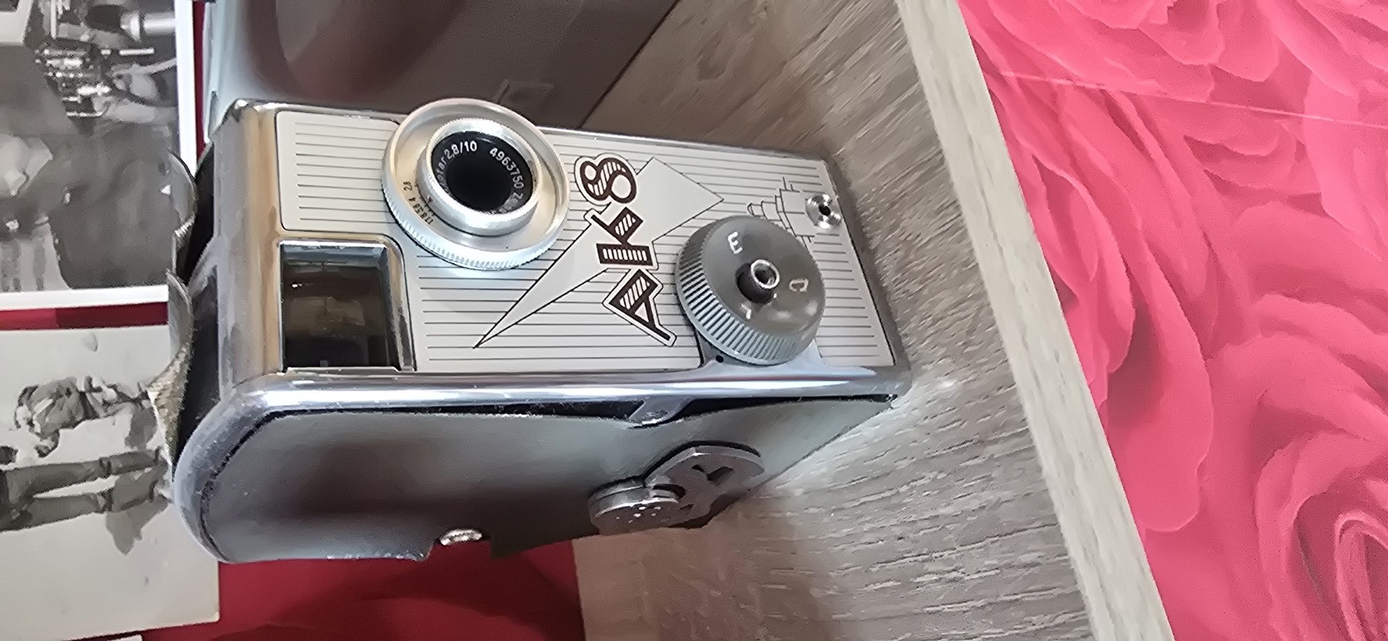 A vintage AK8 camera is displayed on a wooden shelf. The camera has a metallic body with a leather case. The lens is visible, along with several dials and a small view finder. The camera is positioned in front of two black and white photographs, one of which depicts two figures standing in snow. The shelf is a light wood grain and it rests on a bright pink, floral-patterned surface. The overall scene is a close-up shot of the camera and its immediate surroundings. The location is unknown and no specific details about Gostagayevskaya, Russia can be inferred from the image.