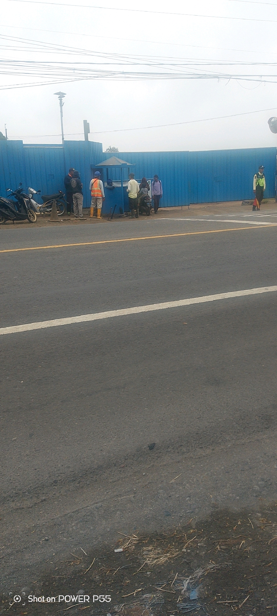 A group of people are gathered near a blue barrier fence with a small guardhouse. Two motorcycles are parked nearby. One person on the right side of the image is wearing a high-visibility vest and appears to be a security guard. The scene is on an overcast day, with power lines visible in the sky. The ground is paved, with a yellow line marking the road.