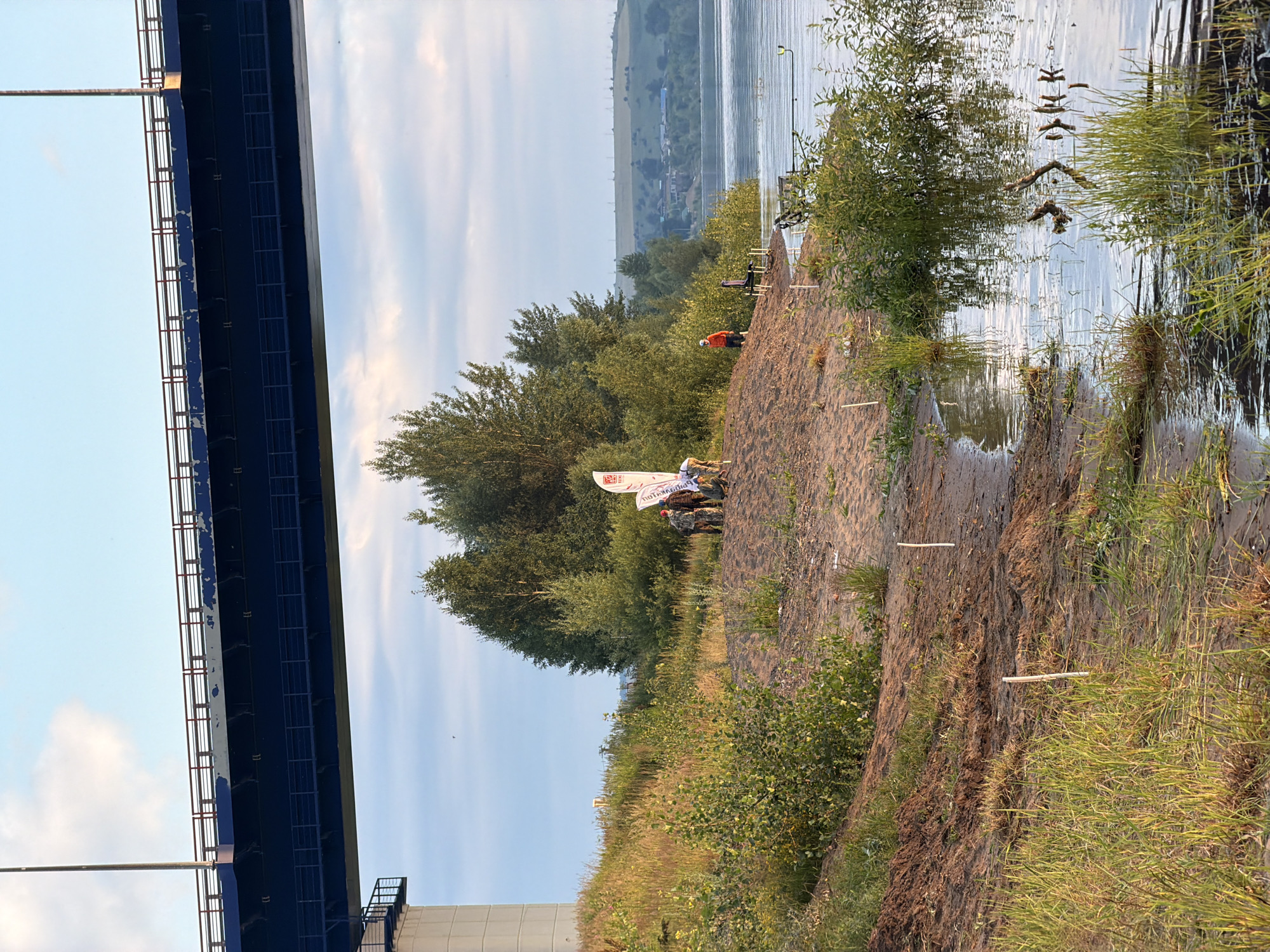 In Kambarka, Russia, under a large blue bridge, a group of people are gathered on a sandy bank next to a river. A white flag with red writing is planted in the sand. One person in an orange jacket stands further back. There are fishing rods set up near the water's edge. The river reflects the sky, and trees line the opposite bank, with a small village visible in the distance. The ground near the river is muddy with tall green grass.