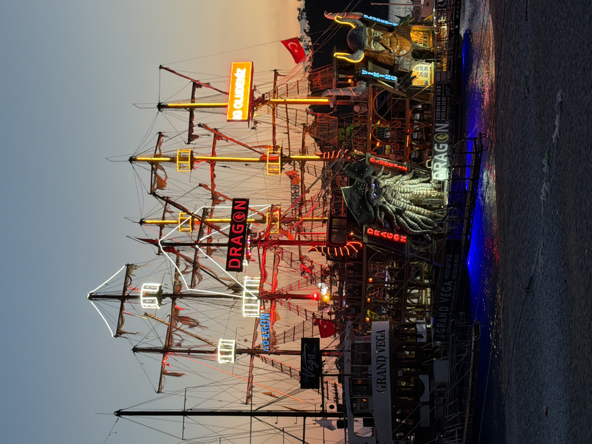 The image shows a nighttime view of decorated ships in Fethiye, Turkey. The ships, heavily adorned with lights, signage, and themed decorations, are likely docked in a harbor or marina, with their masts and rigging prominently displayed. The neon signs read "DRAGON," "IG OLUDENIZ," and "GRAND VEGA." A dragon sculpture, along with other themed figures like a Viking, is mounted on one of the ships. The Turkish flag can be seen on one of the ships. The water reflects the colorful lights of the ships.