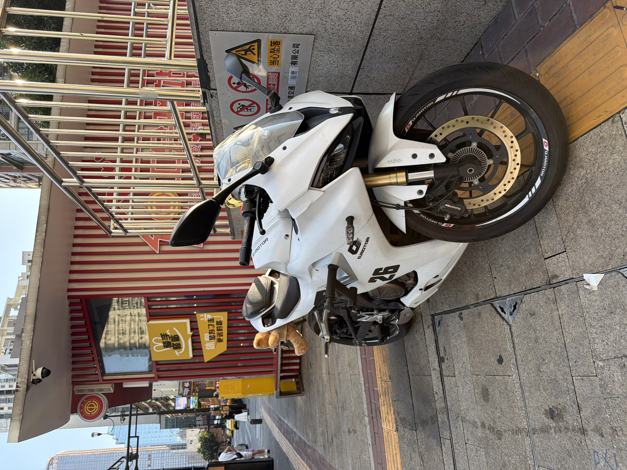The image shows a white sport motorcycle parked on a sidewalk in Yuzhong District, China. The motorcycle is positioned at an angle, with its front wheel on a tactile paving. A teddy bear is attached to the rear seat of the motorcycle. Behind the motorcycle is a red and white striped building with signage in Chinese characters. The building appears to be a small commercial establishment, possibly a food vendor, as indicated by yellow signage with cartoon rabbits. A metal railing runs along the top of the sidewalk and over a set of stairs. In the background, there are modern buildings, a street with traffic, and a few pedestrians. The sky is clear and the lighting suggests it is daytime. A warning sign with pictograms and Chinese text is affixed to the wall behind the motorcycle.