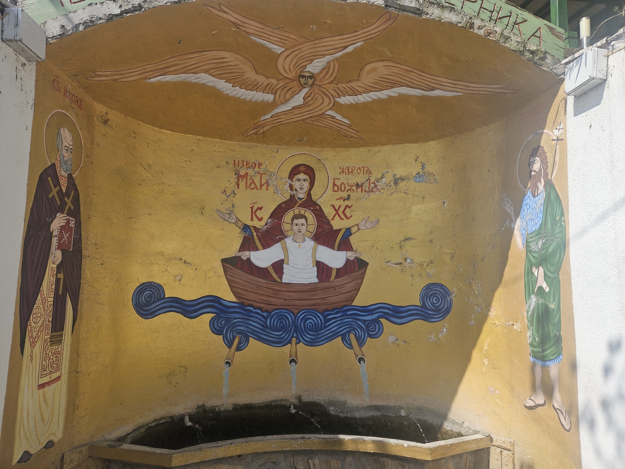 This media file displays a vibrant Orthodox Christian mural painted on a curved, golden-yellow wall, functioning as a fountain. The artwork features several religious figures and iconography, typical of the "Spring of Life" (Životvorni Istočnik) theme.
At the top center, a celestial being with a human face and six broad wings, possibly a seraph or cherub, is depicted. Below, in the central section, the Virgin Mary (Theotokos), wearing a red and blue robe with a halo, extends her arms in an orans pose. She holds the Christ Child, also with a halo and wearing a white tunic, who stands in a wooden boat. The boat rests on stylized blue wavy water, from which three spouts project, channeling water into a stone basin below.
Flanking the central scene are two saints. On the left, a bearded male saint with a halo, dressed in dark bishop's vestments (possibly a phelonion and omophorion with crosses), holds a red book. Text above him identifies him as "СТ НИКОЛА" (Saint Nicholas). On the right, another bearded male saint with a halo, wearing a green tunic and blue undergarment, holds a staff topped with a cross. This is likely Saint John the Baptist.
Visible text above the central figures reads "ИЗВОР ЖИВОТА" (Spring of Life) on the left and "БОЖИЈА" (God's/Divine) on the right, referring to "God's Spring of Life." Below these, "МАЈК" (Mother) and "ХС" (initialism for Jesus Christ) are present. A partial word, "РНИКА", is visible at the top right, possibly part of "ВОДОПРЕНИКА" (water spring/fountain).
The mural is illuminated by sunlight, casting shadows on the right side. The paint shows signs of weathering, with cracks and fading visible on the golden background and some figures. The water from the spouts flows into a dark stone trough, confirming its use as a fountain. The overall scene suggests a holy well or shrine.