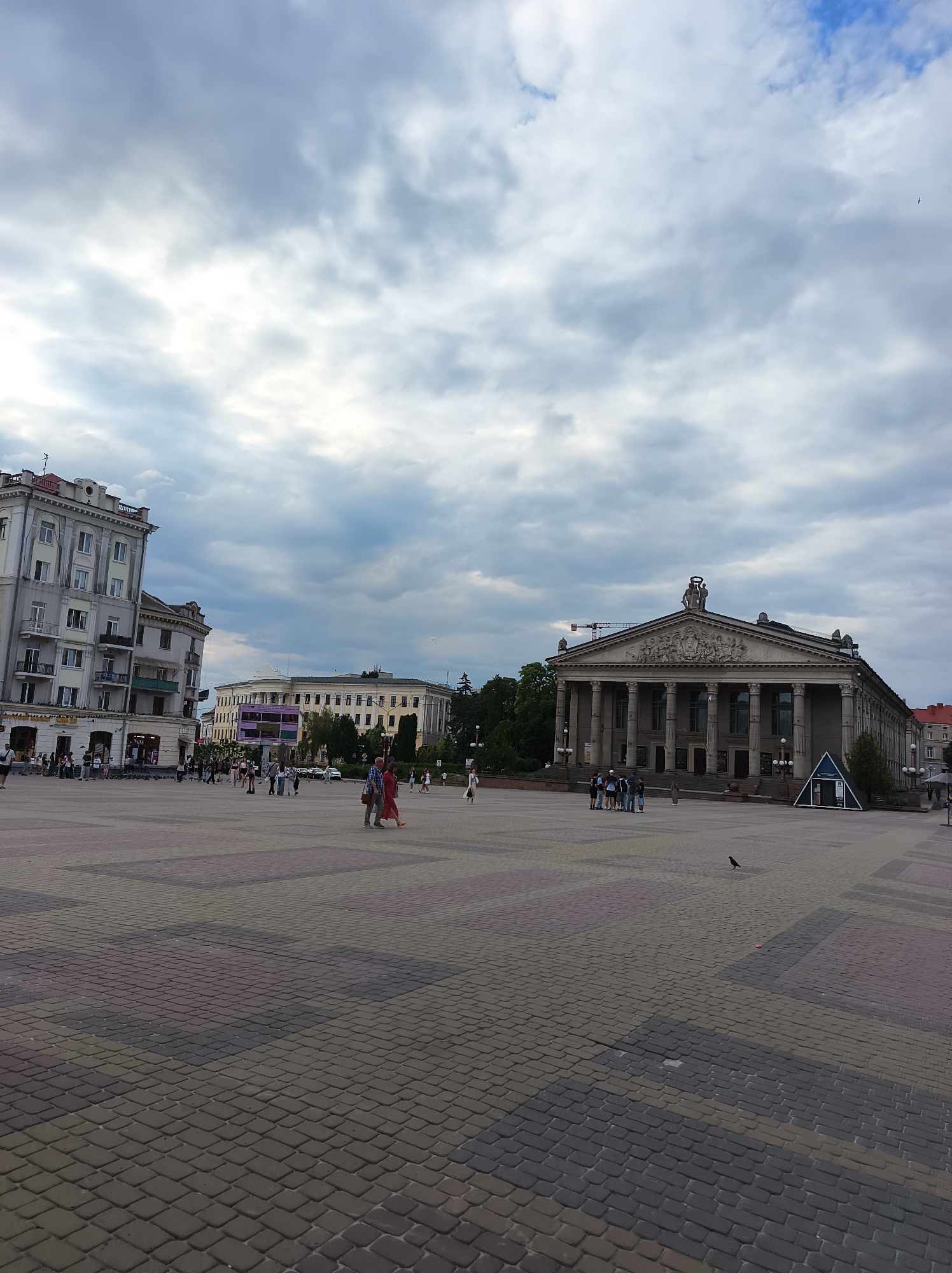 The image captures a wide view of Ternopil's Teatralna Square. In the foreground, the square is paved with a patterned arrangement of dark grey and reddish-brown rectangular stones. A solitary black bird stands on the pavement to the right of the center. In the middle ground, several people are walking across the square. To the left, a multi-story building with a light-colored facade, featuring numerous windows and balconies, dominates the scene. Further to the right, a grand building with a classical facade, characterized by a colonnade and ornate sculptural details above the entrance, is visible. This building appears to be a theater or cultural center. In the background, more buildings, trees, and a cloudy sky complete the urban landscape. The overall atmosphere suggests a public gathering space in a Ukrainian city.