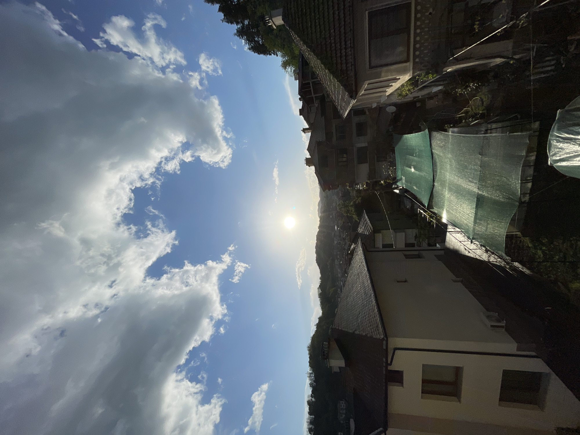 The image depicts a daytime view in Zlatograd, Bulgaria. The sky occupies the left half of the frame, featuring a clear blue expanse interspersed with puffy white clouds. The sun is positioned centrally, radiating a bright glare. The right half of the frame showcases residential buildings, predominantly constructed with stone or concrete. One building has a roof with red tiles. Green netting partially obscures a section of the buildings. A hillside covered in trees descends from the upper center of the frame towards the buildings.