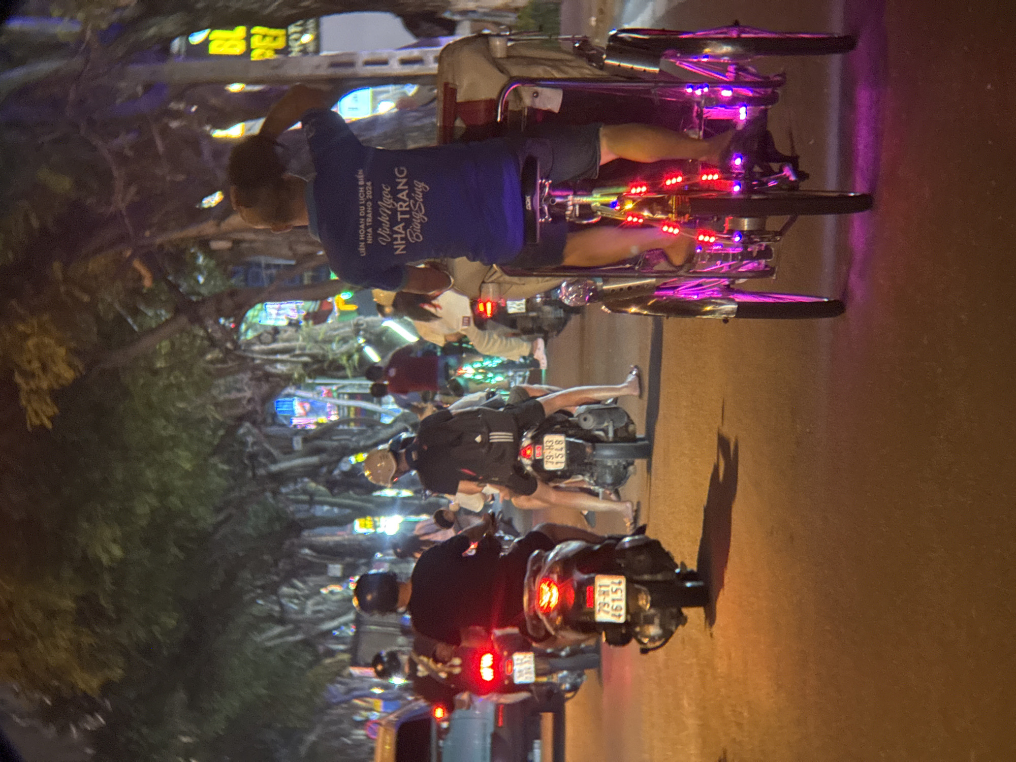 The image captures a street scene in Nha Trang, Vietnam, at night, likely on a coastal road due to the presence of trees commonly found along such routes and the general ambiance. The focus is on a brightly lit cyclerickshaw, a three-wheeled vehicle, decorated with numerous colorful LED lights in red and purple, traversing the street. A person is seated on the cyclerickshaw, wearing a dark blue t-shirt with Vietnamese text that appears to be a slogan related to tourism and "Nha Trang 2024." Several motorbikes are also visible on the road, some with illuminated taillights and license plates. People are riding these motorbikes, some wearing helmets. The street is asphalt, and the surrounding environment is characterized by trees with dense foliage, suggesting a well-treed urban area. The lighting suggests artificial illumination from streetlights and possibly signs from nearby establishments, contributing to the vibrant, nocturnal atmosphere of a tourist destination.