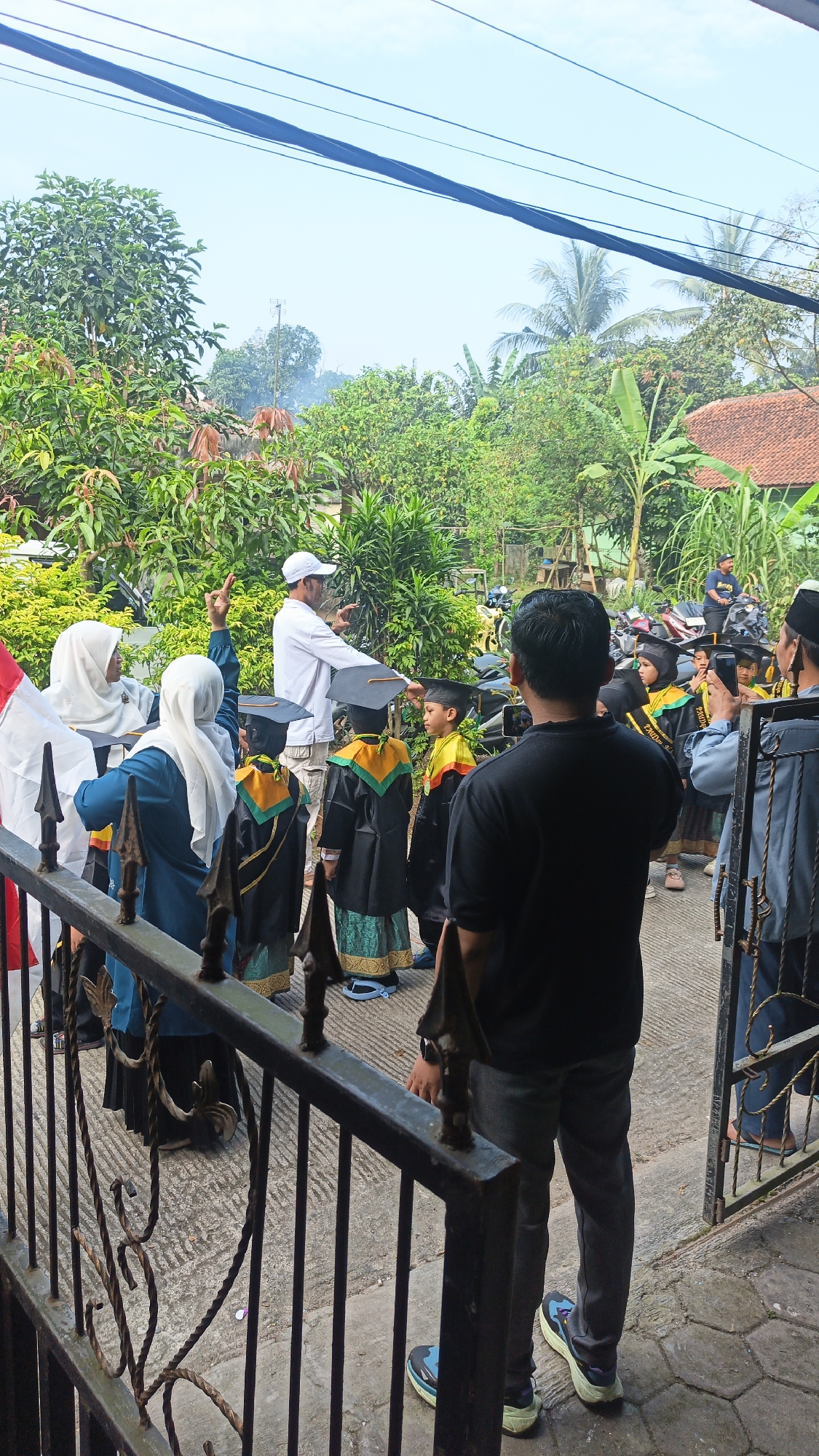 In Tigaraksa, Indonesia, a group of children dressed in graduation gowns and caps are gathered outdoors. Some of the children are holding graduation scrolls. A man wearing a white shirt and a white cap appears to be directing the children. A woman in a blue hijab and a blue dress is also present, standing near the children. Another woman in a white hijab and a blue outfit is also visible. The ground is paved with concrete and there are some metal railings in the foreground. Behind the group, there is lush green vegetation and some houses with tiled roofs. Several motorcycles are parked in the background. The overall scene suggests a graduation ceremony or celebration.