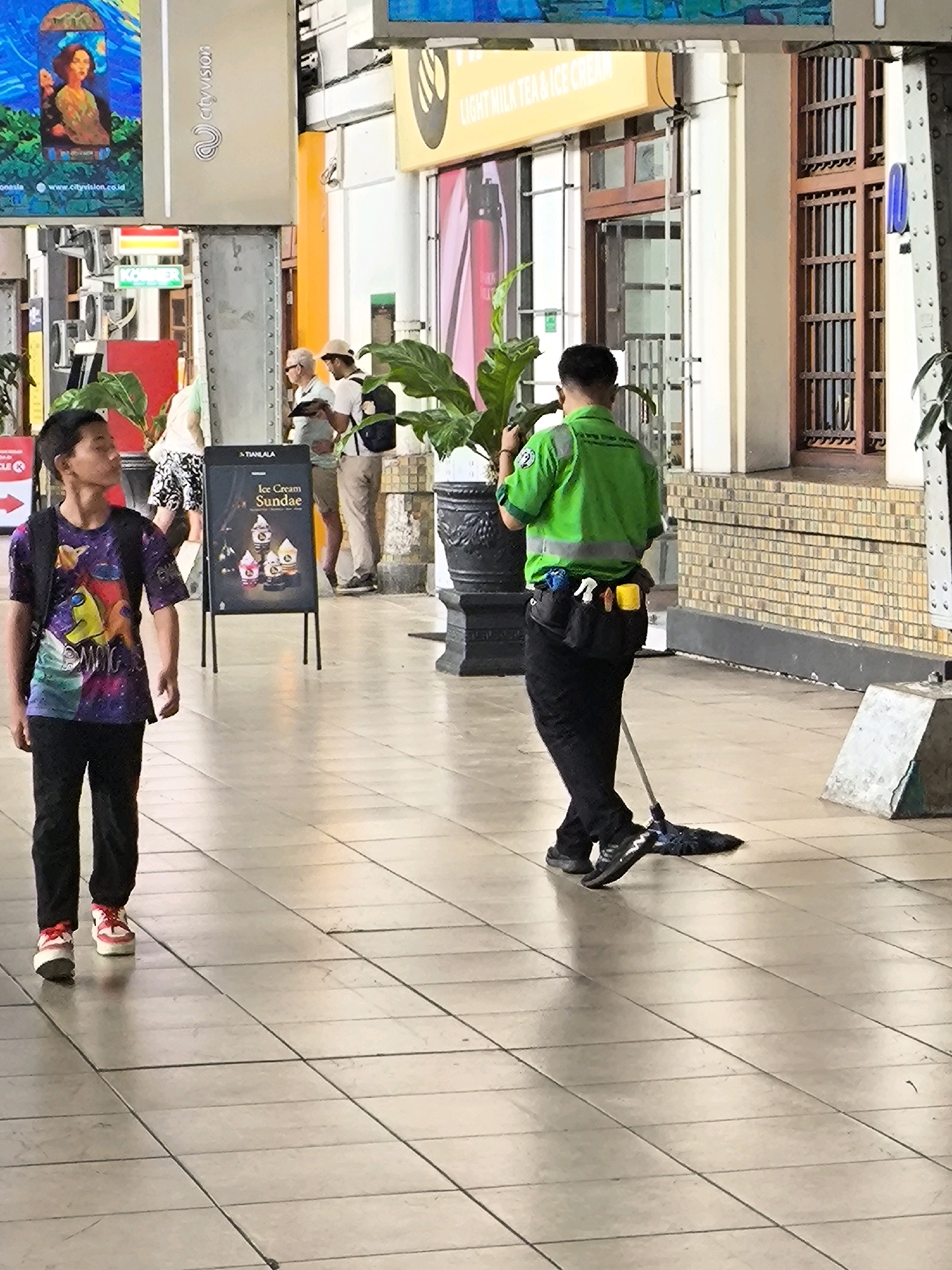 In a covered walkway in West Jakarta, Indonesia, a young boy wearing a galaxy-themed t-shirt and black pants walks to the left, looking away from the camera. Behind him, a cleaner in a bright green reflective vest and black pants mops the tiled floor with a blue mop. The cleaner's utility belt holds various cleaning supplies. In the background, a sign advertises "Ice Cream Sundae." Other individuals are partially visible, including a woman with a floral skirt and a man in a light-colored shirt and hat. The environment appears to be an indoor public space with tiled flooring and a brick wall on the right.