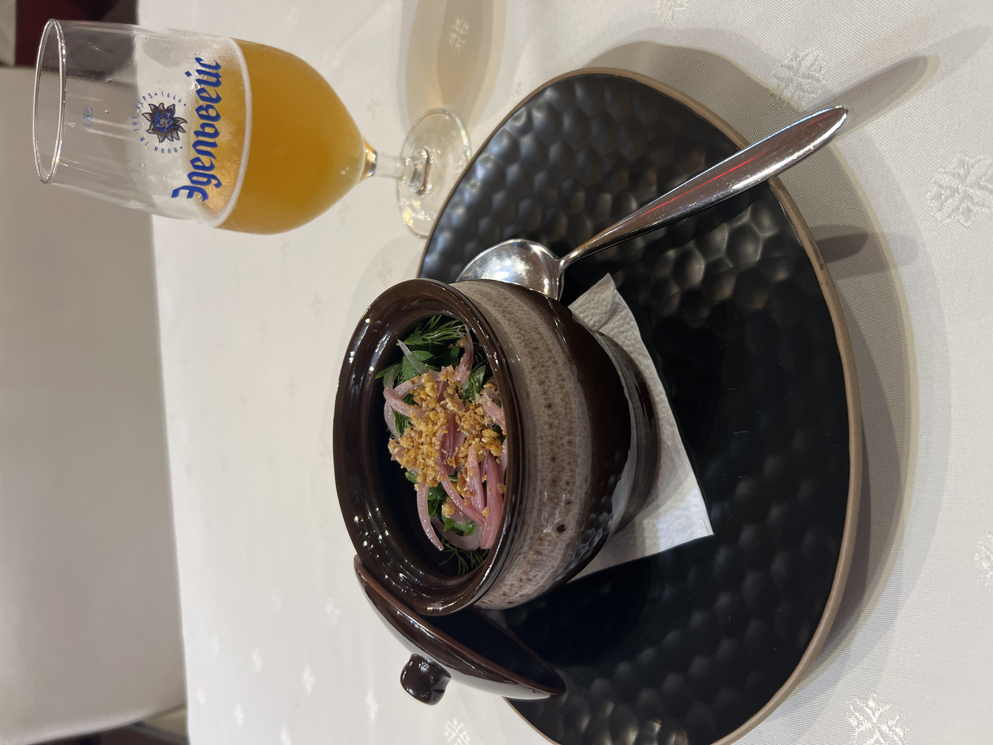 A still life photograph captures a serving of food and a beverage on a white tablecloth. In the foreground, a dark brown ceramic pot with a lighter speckled pattern, partially open, contains a dish topped with thinly sliced red onions, green herbs, and what appear to be fried garlic pieces. The pot rests on a textured black plate with a brown rim. A silver spoon lies across the plate, pointing towards the bottom right of the frame. To the right of the pot is a tall, clear glass filled with a pale golden beverage, likely beer. The glass has blue lettering and a floral emblem. The background is softly blurred, showing a suggestion of a white chair. The scene is lit, casting subtle shadows from the objects onto the tablecloth. The location is unknown.