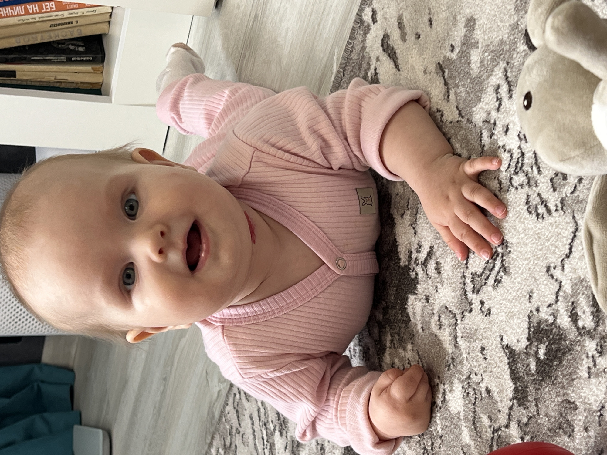 An infant is seen lying on its stomach on a light-colored, patterned rug in an indoor setting in Chaikovsky, Russia. The baby, with light hair and blue-grey eyes, is wearing a pink ribbed long-sleeved onesie or top. Its mouth is open in a joyful or surprised expression, and its eyes are wide and focused, suggesting curiosity or engagement with something out of frame or the camera. A small, light brown patch with an animal silhouette (possibly a rabbit or bear) is visible on the side of the onesie. A reddish smudge is noticeable on the baby's neck area, possibly lipstick or a mark.
To the baby's right, a light grey stuffed animal, appearing to be a rabbit or similar creature, is partially visible. In the background, to the left of the baby, a stack of books rests on a white shelf. Visible titles on the books include "БЕГ НА ЛИНИИ" (Run on the Line), "Джеки Чан" (Jackie Chan), "КРИМИНОЛОГИЯ" (Criminology), "BACKTRACK," and "Basketball," indicating a mixture of Russian and English titles. The floor beneath the rug appears to be a light wood or laminate. The overall lighting suggests it is daytime. The baby's posture and expression convey a happy and active moment of "tummy time."