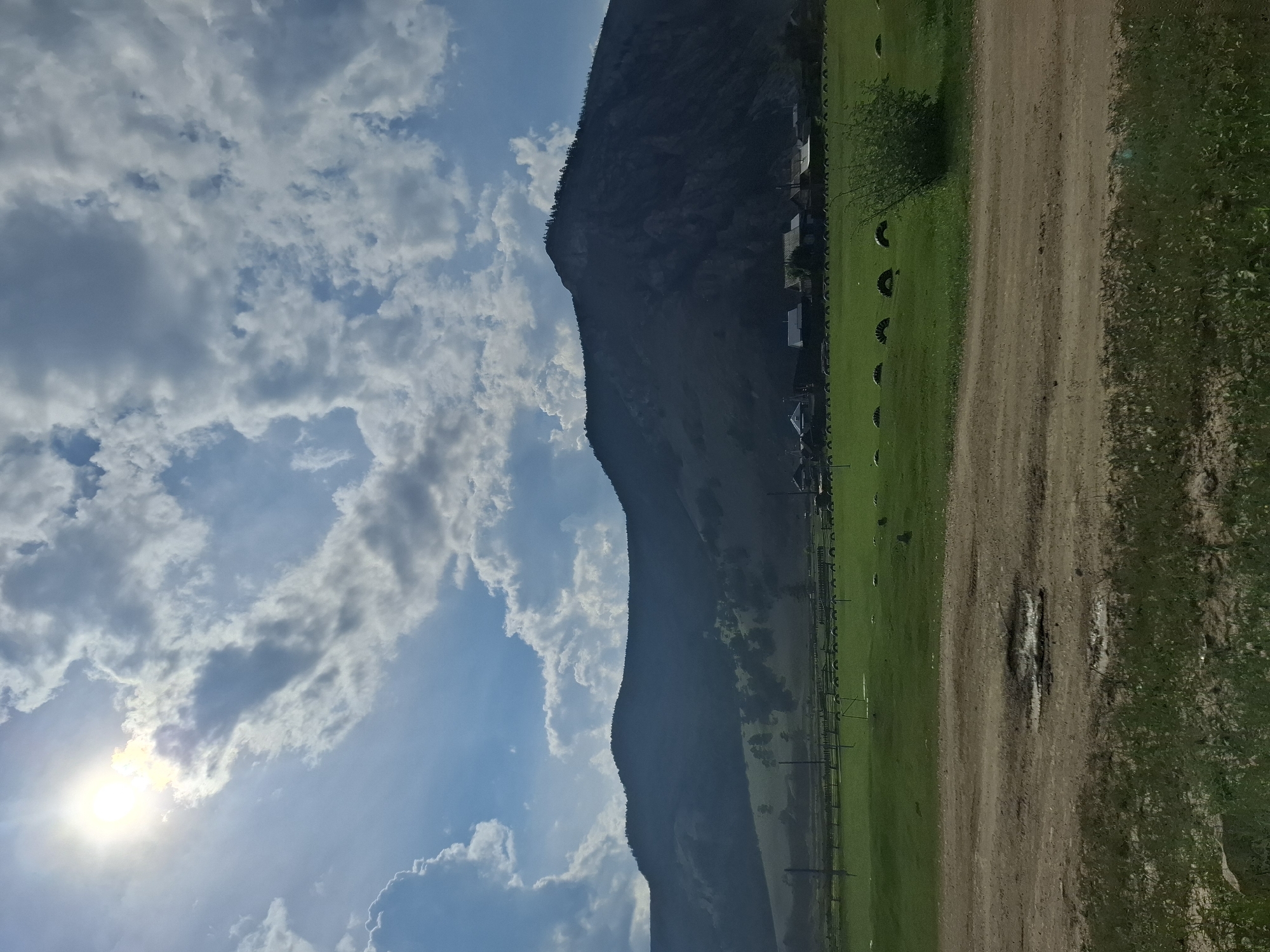 The image depicts a rural landscape under a partly cloudy sky. In the foreground, a dirt road with puddles is visible. Beyond the road, a vast green field stretches towards a line of houses and a dark, imposing mountain range in the background. The sun, partially obscured by clouds, shines brightly in the upper left corner. The scene appears to be located in Buguldeyka, Russia, a village situated on the western shore of Lake Baikal, known for its scenic natural beauty.