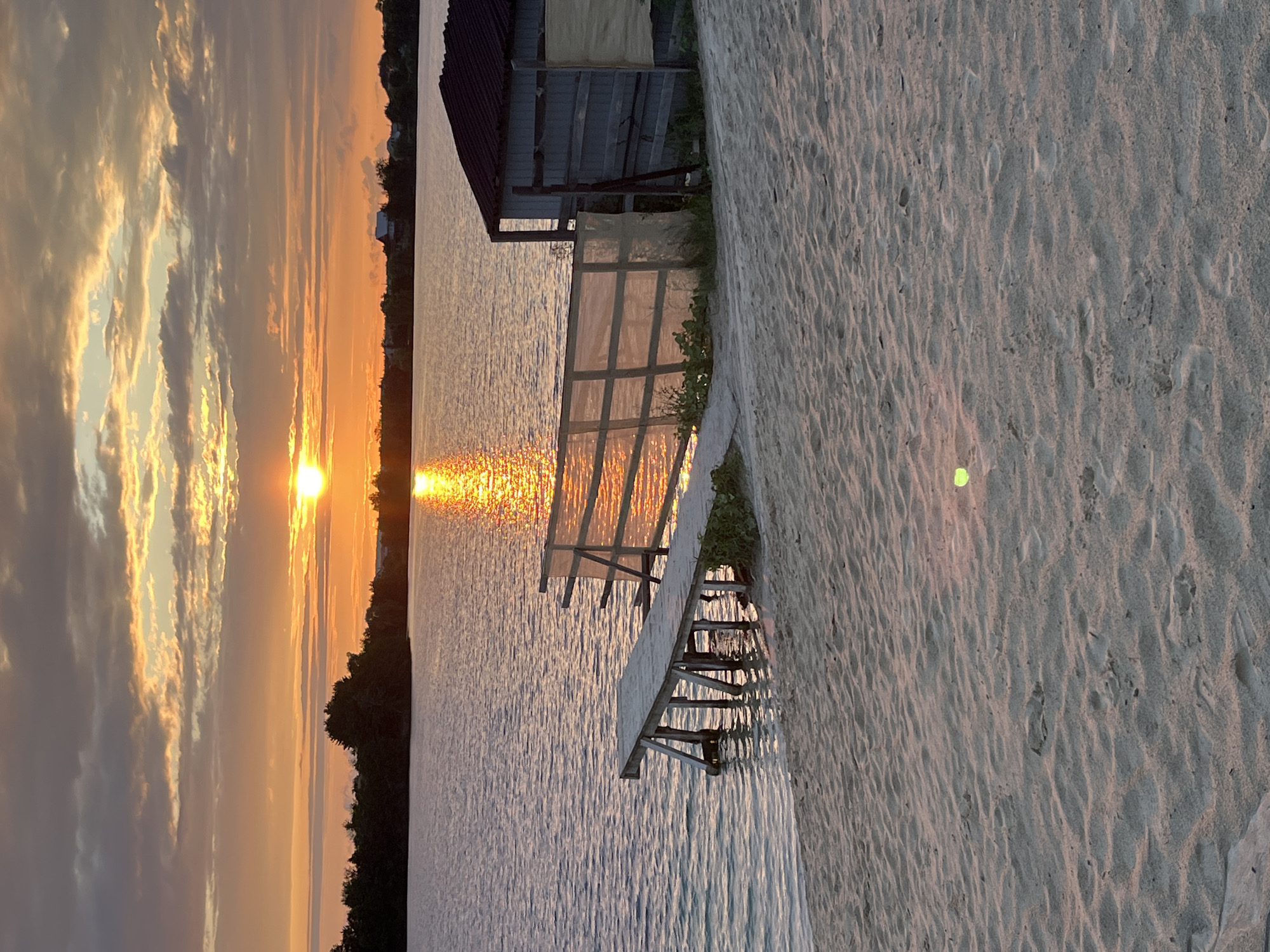 The image depicts a sunset over a body of water, likely a lake or reservoir, in Bezliudivka, Ukraine. The foreground features a sandy beach with faint footprints. Extending into the water is a wooden pier with visible supports. Adjacent to the pier, on the sandy bank, is a small, rustic wooden structure with a dark, sloped roof and grey wooden walls. A translucent fence-like barrier is positioned between the pier and the structure. The water reflects the warm hues of the setting sun, creating a shimmering golden path. The horizon is lined with dark trees and some distant buildings. The sky is filled with dramatic clouds, tinged with orange and yellow from the setting sun. A small, bright green object, possibly a tennis ball, is visible on the sand in the lower right quadrant of the image. The scene is tranquil and serene, capturing the natural beauty of the location at dusk.