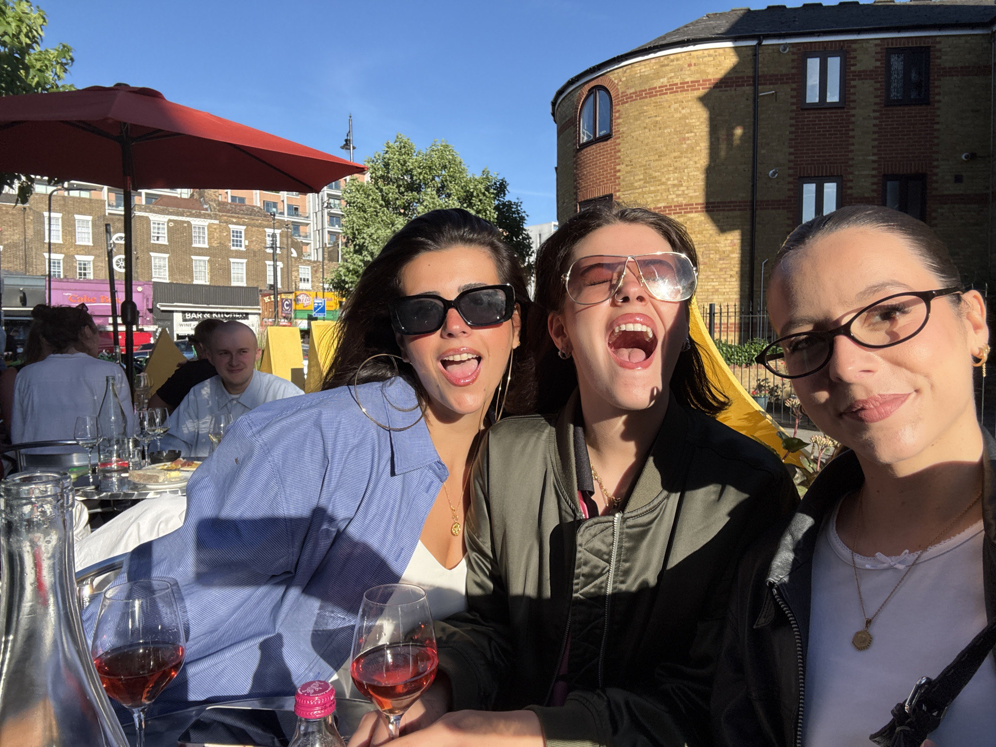 The image shows a group of people sitting at an outdoor table, possibly a cafe or restaurant, with the London skyline visible in the background. Three young women are in the foreground, one on the left wearing a blue shirt and large sunglasses, one in the middle wearing a green bomber jacket and aviator sunglasses, and one on the right wearing glasses and a white top. They are all smiling and appear to be enjoying themselves. There are drinks and food on the table, including glasses of rose wine and a bottle of wine. The setting appears to be a sunny day in London, with buildings and trees visible in the background.