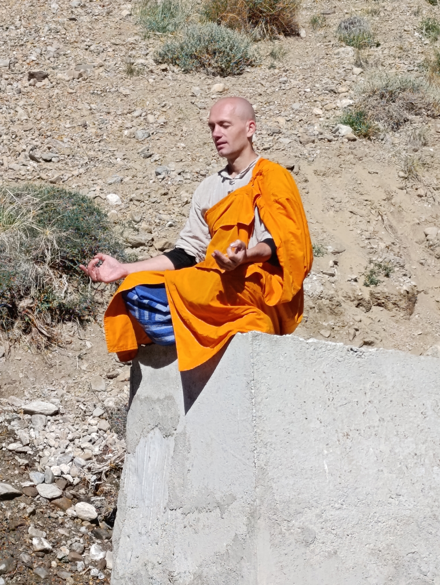 A bald man wearing an orange robe and a light-colored shirt sits in a meditative pose on a concrete structure. He is outdoors with a rocky, barren hillside behind him, dotted with sparse vegetation. His eyes are closed, and his hands are held in a mudra. The man is in the city of Dhar Chharp Samba, India.