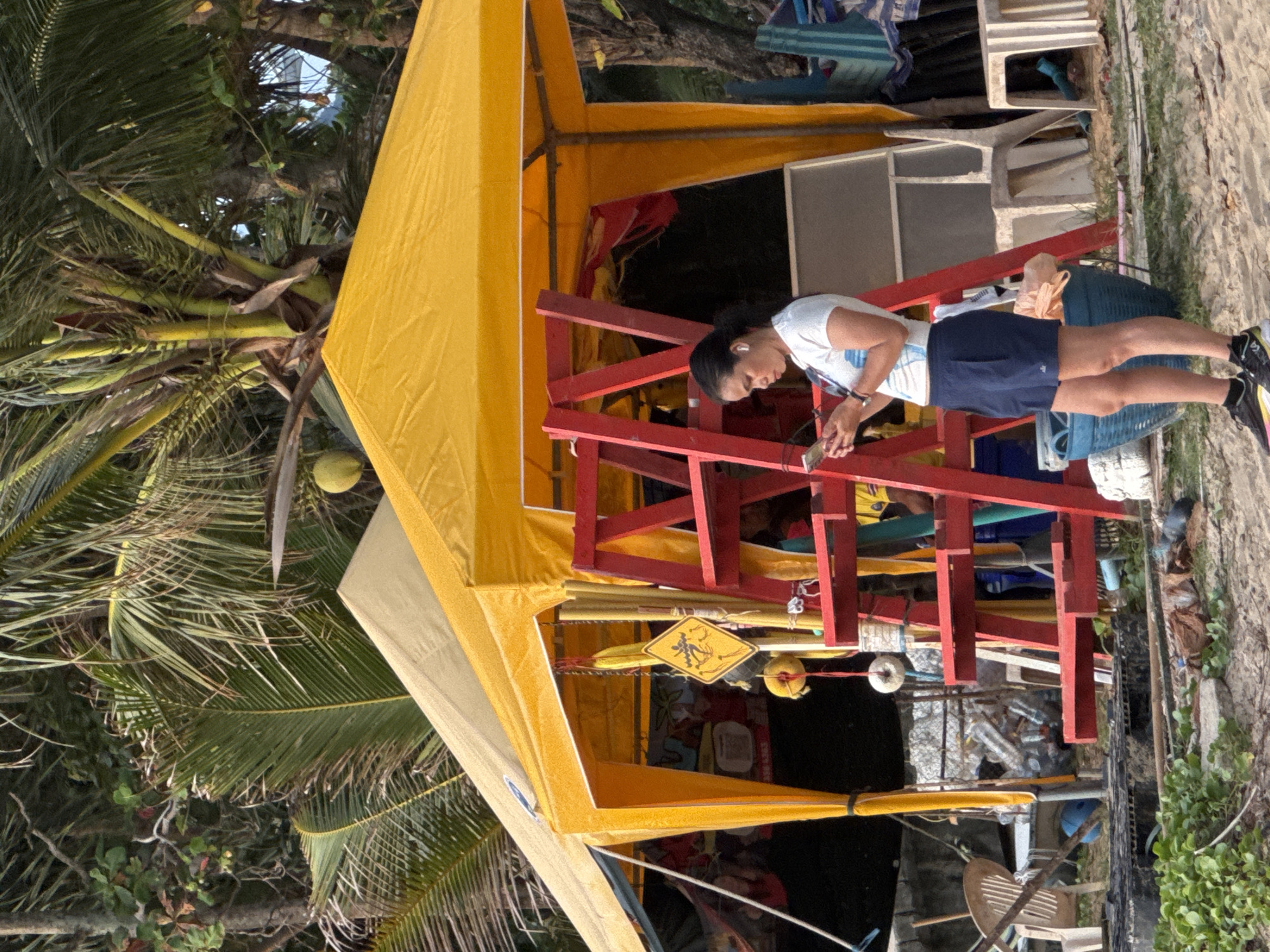 A woman with dark hair tied back in a ponytail, wearing a white t-shirt, navy shorts, and black and yellow athletic shoes, stands on a sandy beach in Karon, Thailand. She is looking down at her mobile phone, holding it in her left hand. She is wearing white earbuds. Behind her is a large yellow tent with a beige top, supported by poles. A red ladder-like structure is positioned in front of the tent. Several objects are visible hanging from the tent, including a yellow diamond-shaped sign with a depiction of a surfer. Behind the tent, a pile of plastic bottles is visible, along with some chairs and other miscellaneous items. Palm trees and foliage are present in the background, indicating a tropical environment. The scene appears to be outdoors, possibly near a market or beachside stall.