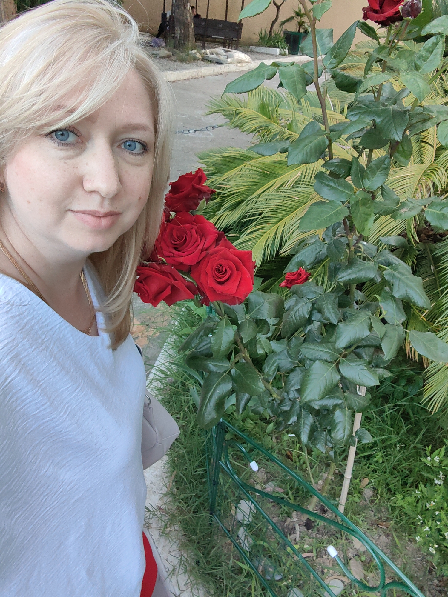 A blonde woman with blue eyes is taking a selfie outdoors, with a rose bush in the background. She is wearing a white shirt with a textured pattern and a gold chain necklace. The rose bush has several red roses. The background shows greenery, a paved walkway, and a fence. The setting appears to be a park or garden area in the city of Sochi, Russia, a popular resort city on the Black Sea coast.