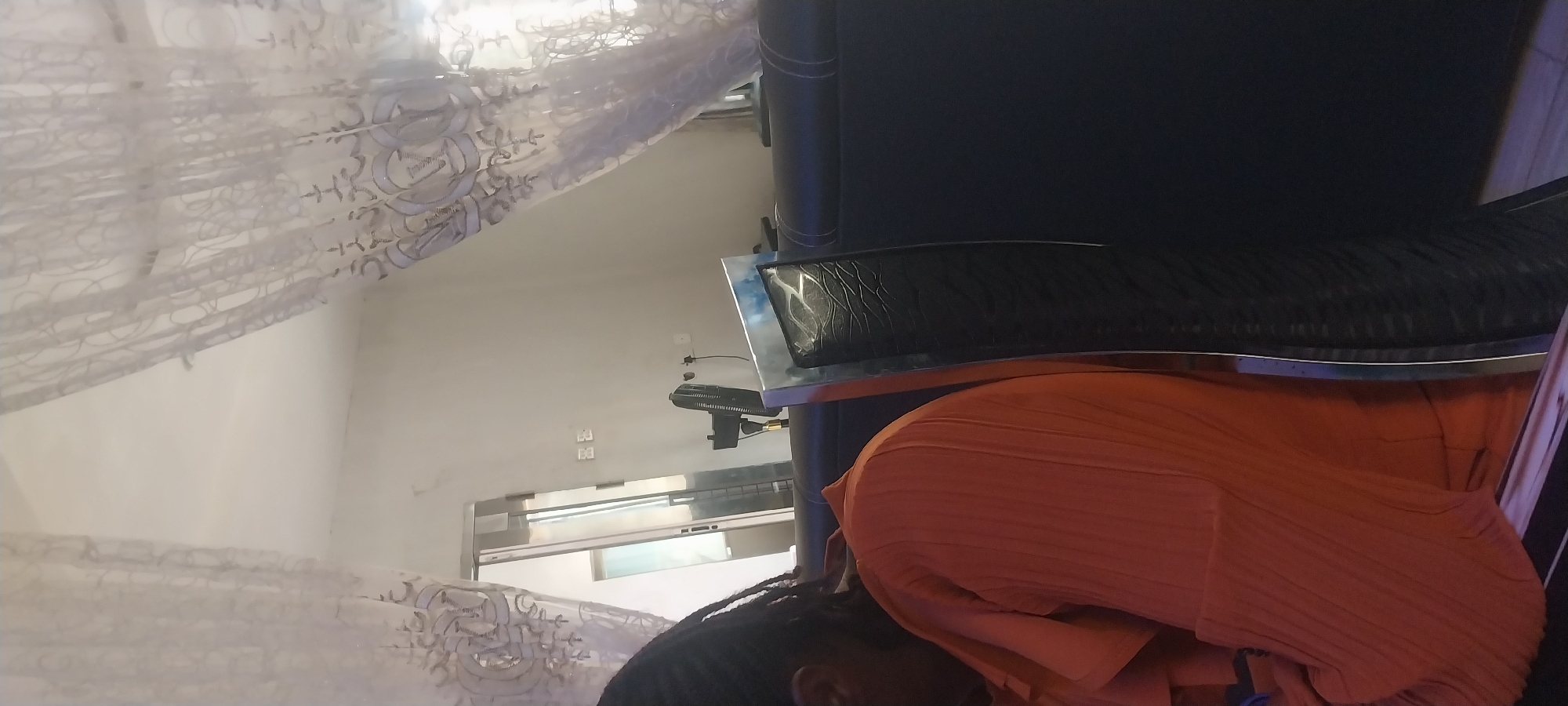 The image shows an interior scene with a person sitting in an orange, ribbed garment. The person is positioned in the foreground, facing away from the camera and slightly to the left. Behind the person, a dark, possibly leather, sofa is visible. To the right of the person is a tall, metallic object with a black textured insert, possibly part of a chair or decorative element. In the background, sheer white curtains with a pattern hang from what appears to be a ceiling or window. Further back, a doorway or opening leads to another room, where a fan is standing. The walls are painted white. The scene appears to be indoors, in a room within a building, possibly a home or a reception area. The location of Akpaka Forest Reserve, Nigeria, is not directly evident from the visual information in the image.