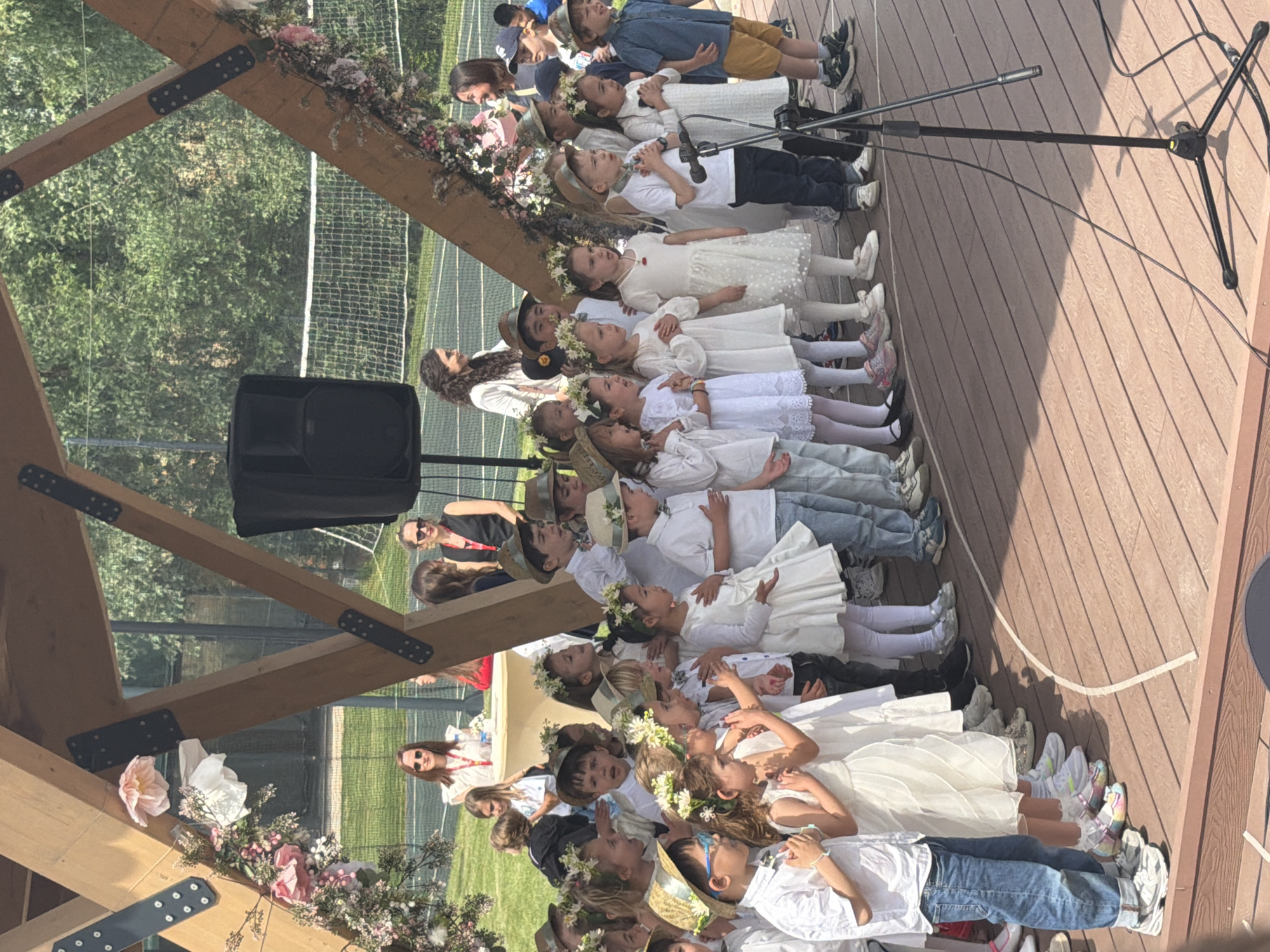 A group of children are standing in a row on a wooden platform, possibly singing. They are a mix of boys and girls, mostly dressed in white with some wearing flower crowns or straw hats. There's a microphone stand in front of them, suggesting a performance. The platform is part of a wooden structure decorated with flowers. Behind them, there is a tennis court and some trees. The location is likely in Cukarica Urban Municipality, Serbia, given the prompt. Adults are visible in the background, possibly supervising or watching the performance. A loudspeaker sits to one side.