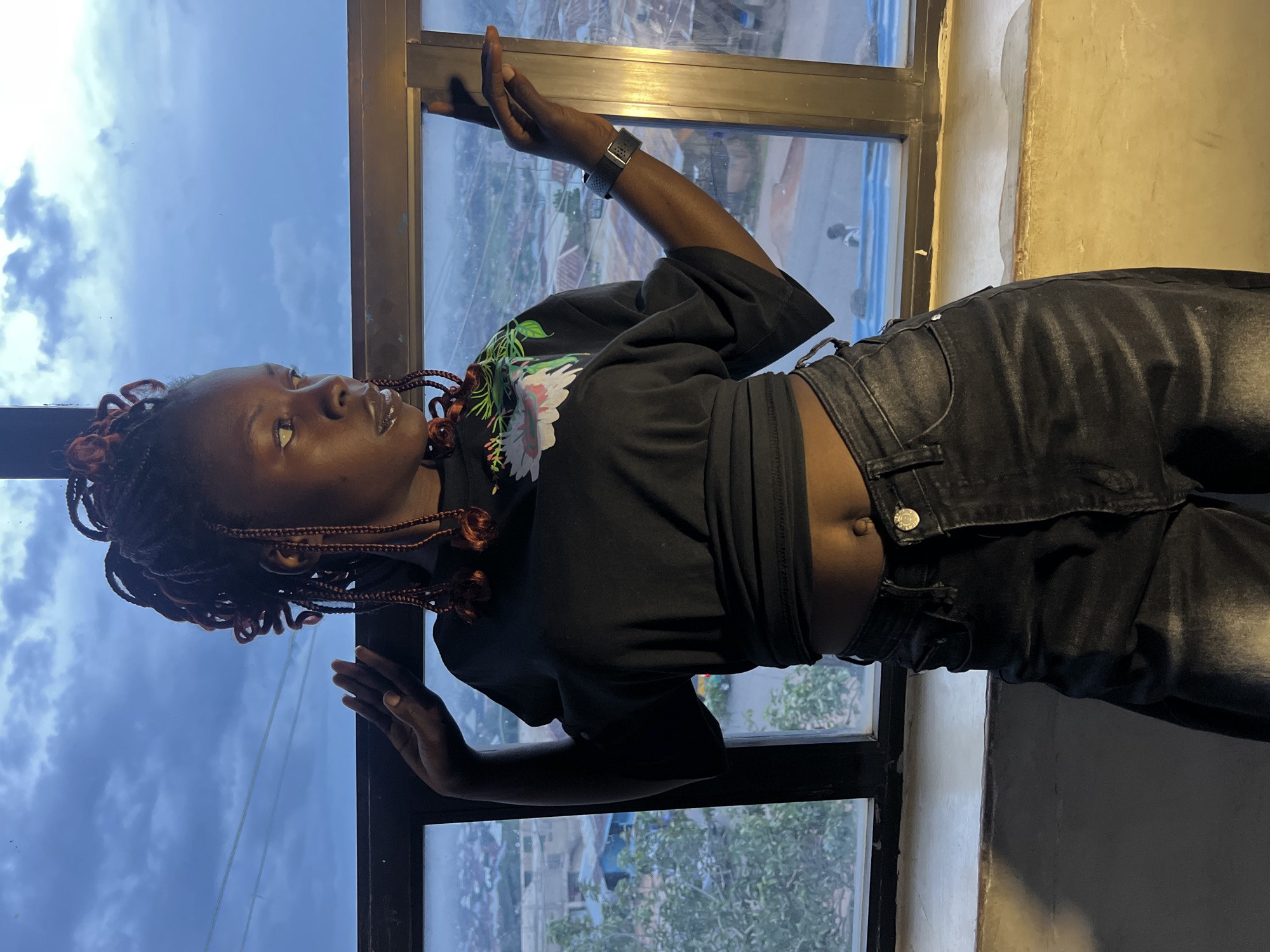 A young Black woman with braided hair, adorned with reddish-brown accents, is positioned in profile against a large window. She is wearing a black cropped t-shirt with a floral design on the left chest and black jeans. Her left hand is raised with fingers extended, and her right hand rests on the window frame. She is looking out at the city of Jos, Nigeria, visible through the window. The sky above is overcast with dramatic clouds. The interior setting appears to be an elevated space, possibly a restaurant or apartment, given the view of the city below. The lighting suggests it is late afternoon or early evening.