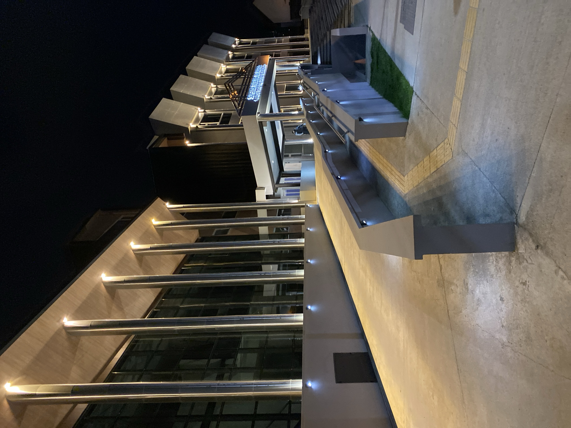This is an exterior shot of the "Superior Tribunal de Justicia" building in Ushuaia, Argentina at night. The building has a modern architectural style with a glass facade, illuminated by horizontal light bars. The building also has cube-shaped structures on its roof. A ramp leads up to the building, lined with illuminated steps and planters. The sidewalk is paved with concrete and tactile paving. The sky is dark.