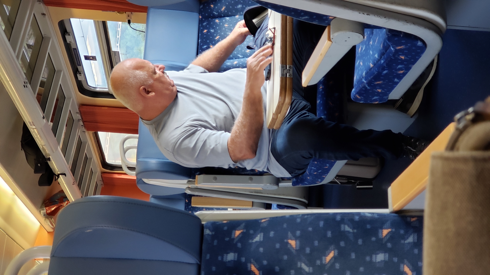 The image depicts the interior of a train carriage, likely in Margecany, Slovakia. A bald man, wearing a grey t-shirt and dark blue jeans, is seated in a blue train seat with a patterned upholstery. He is looking down and appears to be reaching into a black bag placed on a foldable wooden tray table in front of him. A pair of glasses and a mobile phone are also visible on the tray table. The train carriage features overhead storage racks with luggage, and orange curtains are drawn over the windows. The lighting suggests it is daytime.