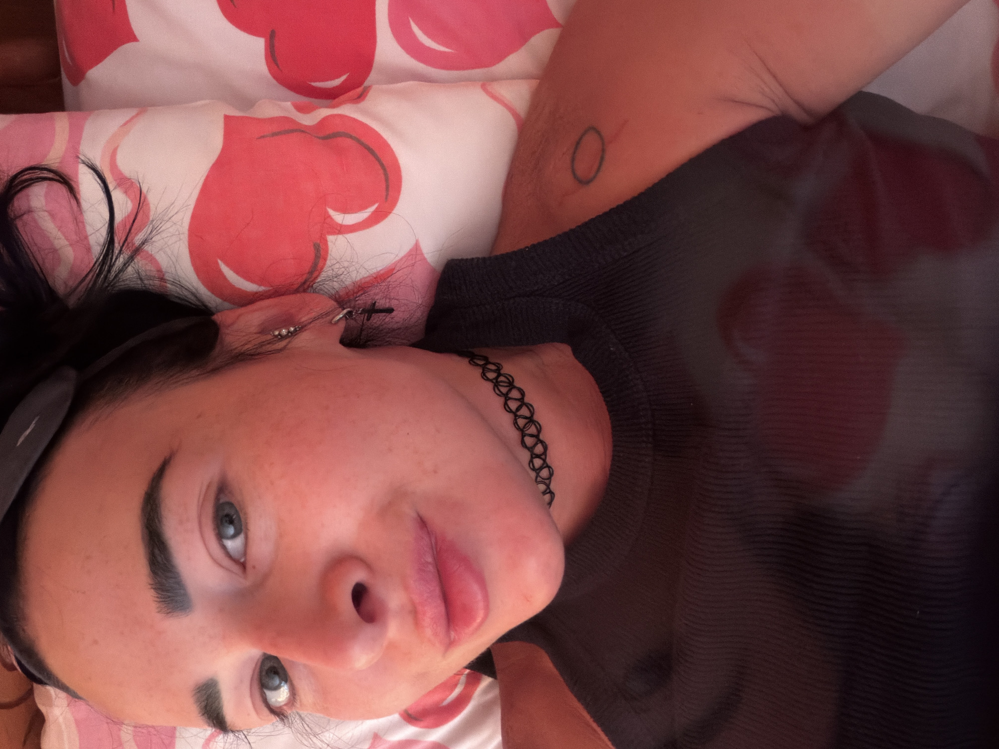 A medium shot shows a woman lying down on a pillow with a red and pink heart pattern, facing the camera. She has dark hair pulled back and is wearing a dark tank top and a black choker necklace. Her right ear has a small silver cross earring. A faint circular tattoo is visible on her upper right arm. The lighting is warm, casting a reddish hue on her skin.