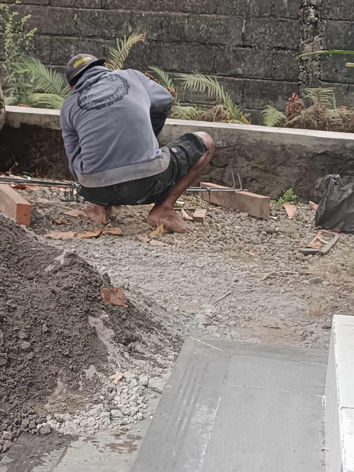 A construction worker, wearing a blue hoodie and dark shorts, squats on the ground in an outdoor setting. The worker has a grey baseball cap on, and is facing away from the camera. In the background, a textured grey brick wall runs horizontally. In front of the wall, there is a low concrete barrier with green foliage growing over it. The ground is covered in small stones and dirt, with a pile of dark soil to the left of the frame. There are also several bricks and some rebar visible on the ground. The scene appears to be in a residential area of Sumberadi, Indonesia.