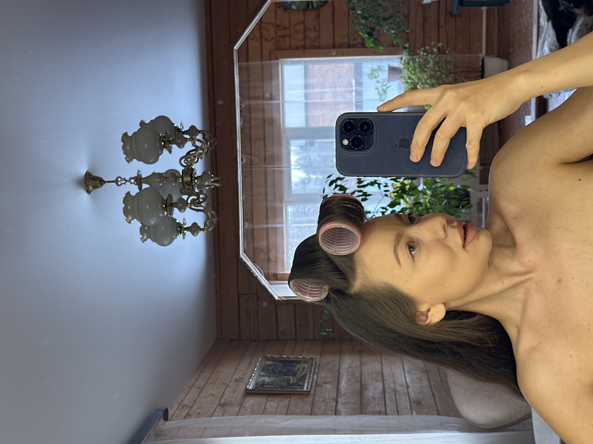 A woman is taking a picture of herself in a mirror. The mirror is in a room with wood paneling, a chandelier, and a painting on the wall. The woman has curlers in her hair and is holding a dark blue smartphone. Outside the window, there appears to be urban structures, potentially locating the scene in a residential area of a city. While the description of the image includes contextual details, there is no explicit information present in the image to confirm the specific location as Kazan, Russia.
