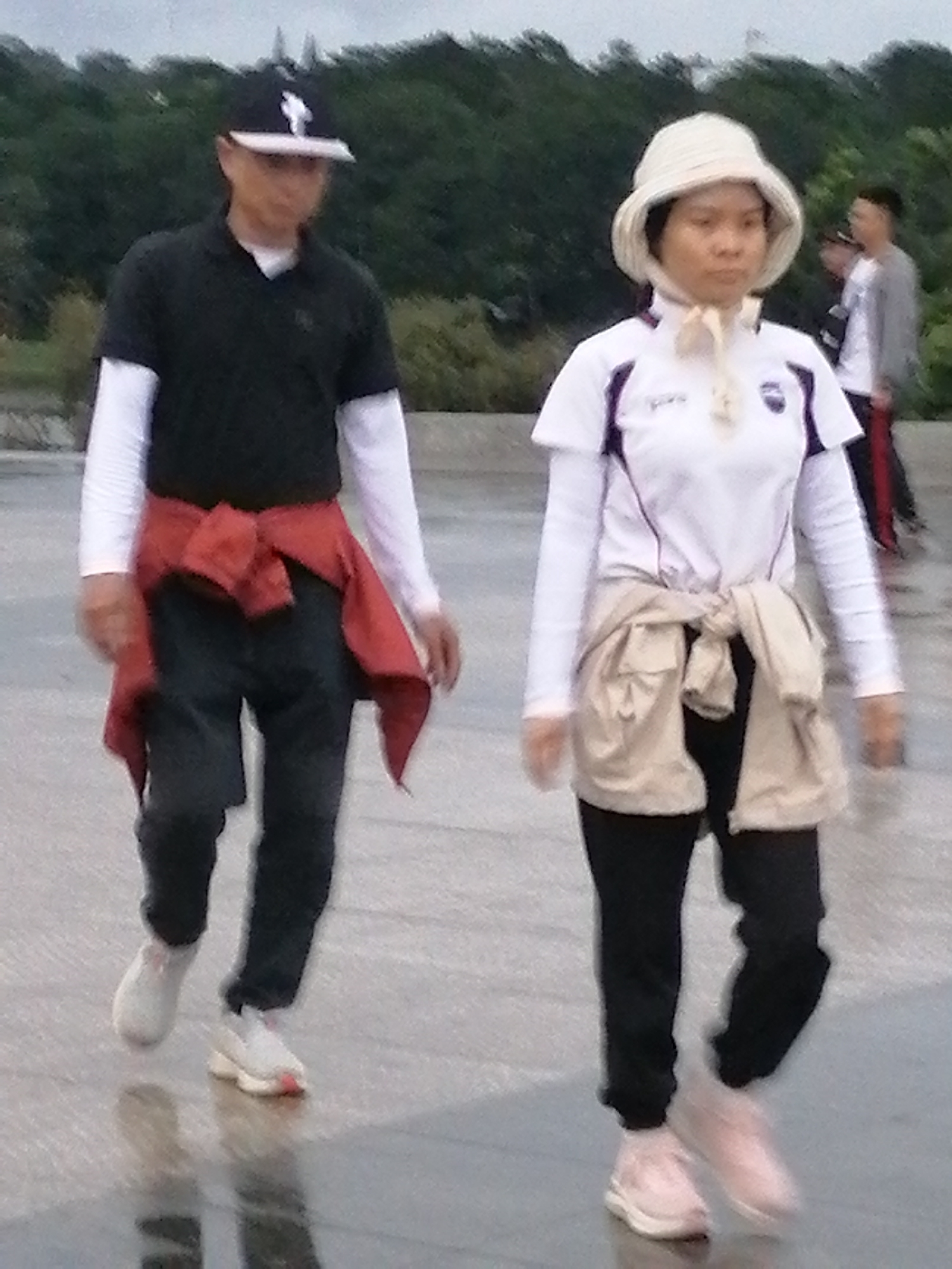 In Da Lat, Vietnam, on a paved, reflective surface that suggests a park or public walkway, two individuals are captured in motion. A man, wearing a black polo shirt with white undersleeves and a black baseball cap with a white emblem, walks to the left of the frame. A red garment is tied around his waist. He wears black pants and white athletic shoes. To his right, a woman, dressed in a white long-sleeved shirt with purple trim and a beige hat, also walks to the left. A light-colored jacket is tied around her waist. She wears black pants and pink athletic shoes. In the background, trees form a dense, dark green backdrop under an overcast sky. Another person, partially visible, walks further in the background to the right, wearing a grey jacket and dark pants. The lighting is diffused, indicating an overcast day.