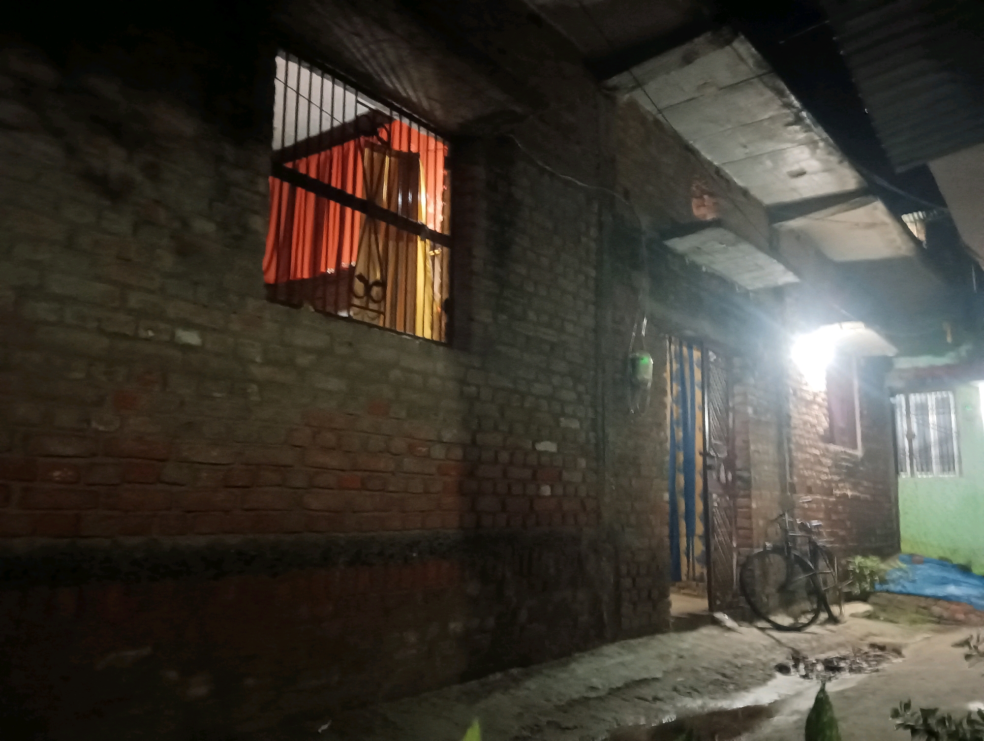 This is an exterior shot taken at night in the city of Dakhin Parsa, India. The scene depicts a narrow alleyway with brick walls and a corrugated metal roof. In the foreground, the ground is wet and appears to be paved with concrete, with some debris scattered about. To the left, a brick wall dominates the frame, with a window that has orange and yellow curtains visible behind metal bars. Further down the alley, a bicycle is leaning against a brick wall next to a doorway. The doorway has a blue and yellow striped door. To the right, another window with metal bars is visible, and part of a green painted wall can be seen. The overall impression is one of a somewhat run-down urban environment at night, with minimal lighting. There are no people visible in the image.