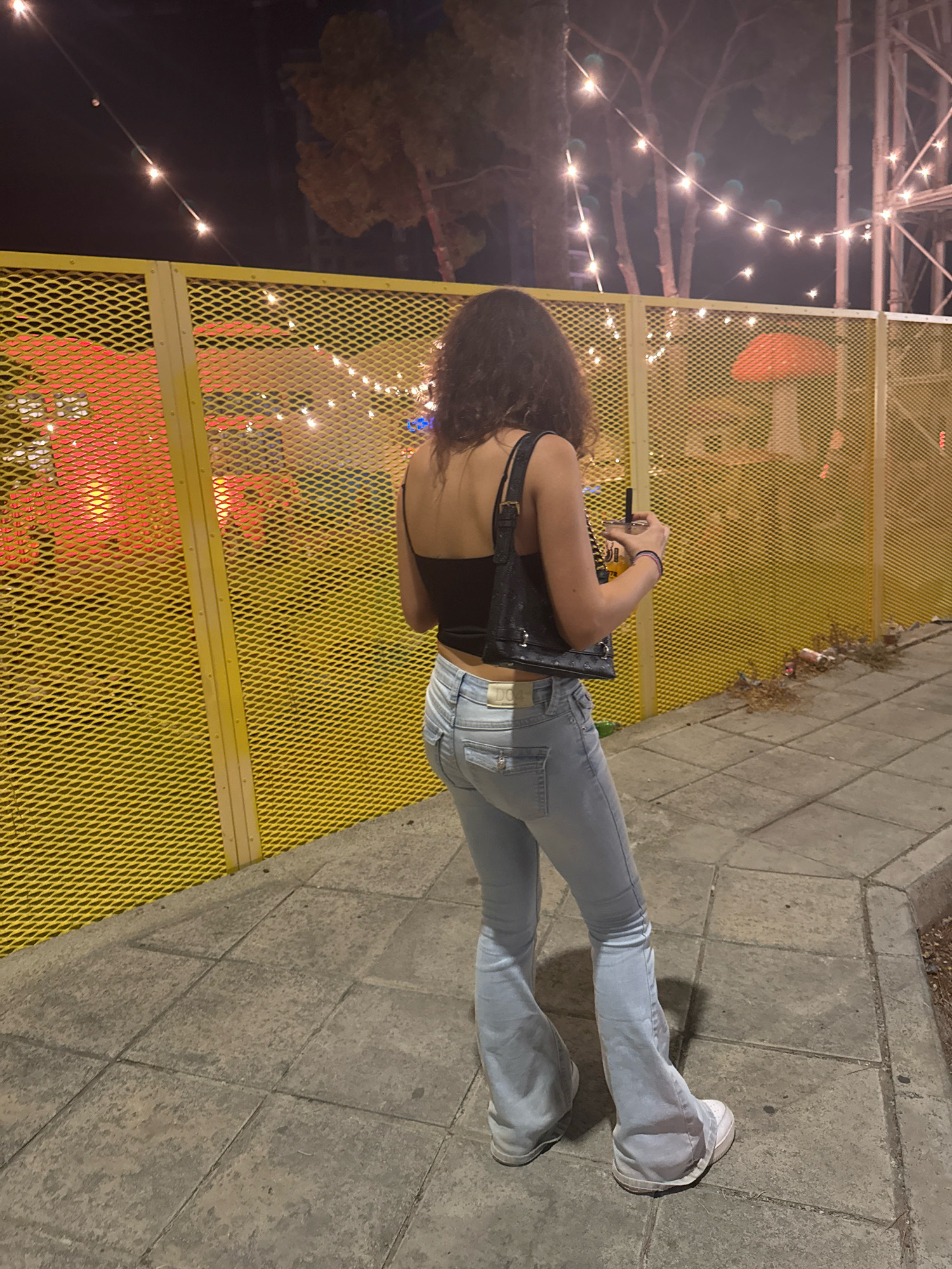 A young woman with dark, curly hair stands facing away from the camera, holding a drink in her right hand. She is wearing a black spaghetti strap top, light-wash flare jeans, and white sneakers. A black crocodile-print handbag is slung over her left shoulder. She is standing on a tiled sidewalk next to a bright yellow chain-link fence. Behind the fence, string lights are visible against a dark sky, and some large, illuminated mushroom-shaped decorations can be seen. The setting appears to be an outdoor venue or event at night. The location is Limassol, Cyprus.