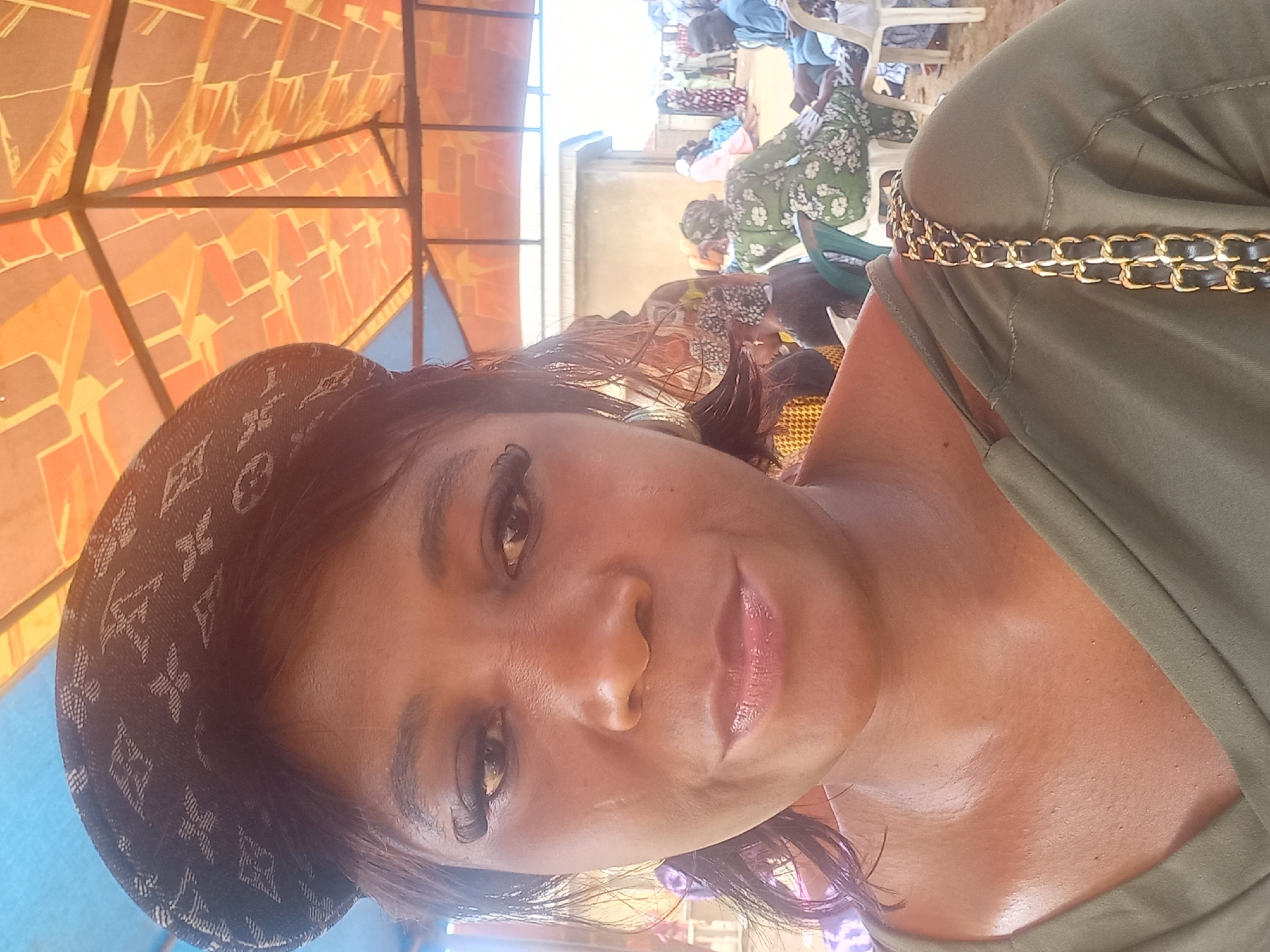 A medium shot shows a woman from the chest up, wearing a dark patterned baseball cap and an olive green off-the-shoulder top with a gold chain strap visible. She has dark hair with some strands falling around her face. She appears to be outdoors, with a colorful, patterned awning visible above her. In the background, several people are seated on chairs, some wearing traditional African clothing. The environment suggests a gathering or event. The image is likely taken in Mangu Town, Nigeria, based on contextual information not present in the image itself.