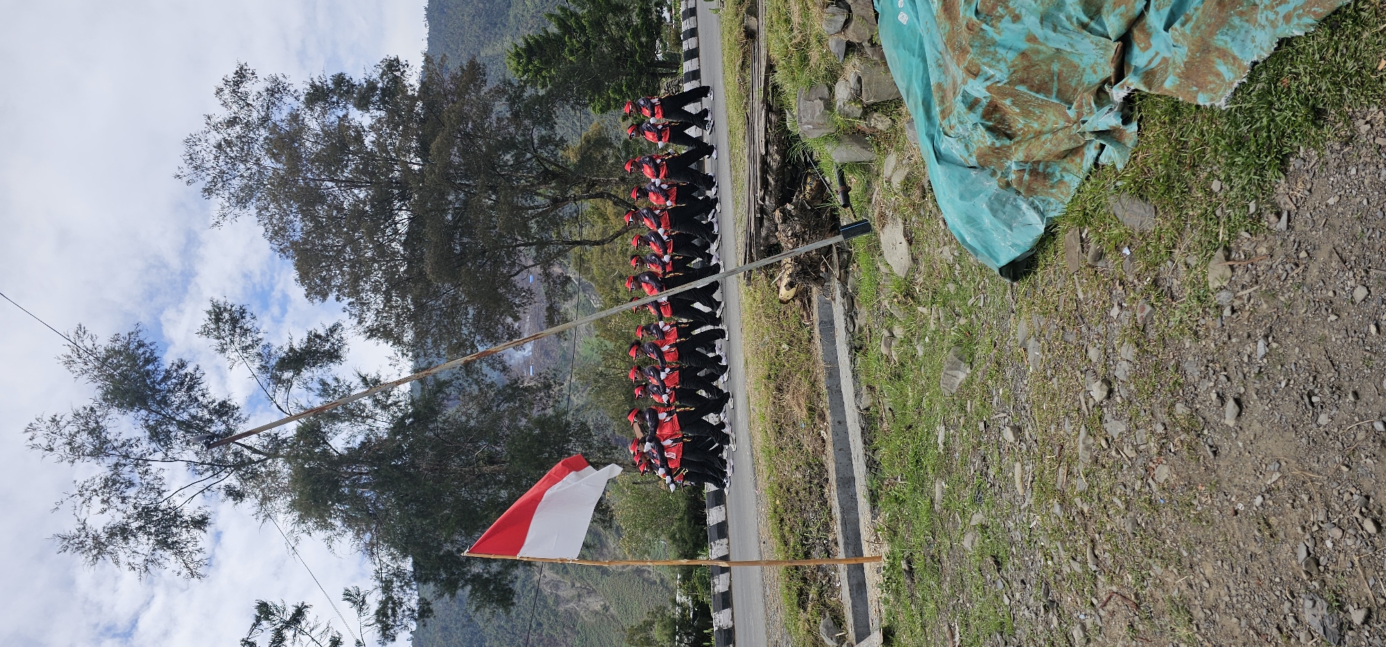 In Puncak Jaya, Indonesia, a group of individuals dressed in red and black uniforms with red caps are marching in formation along a paved road. They appear to be participating in a parade or ceremonial event, as indicated by their synchronized movements and formal attire. A red and white Indonesian flag is prominently displayed on a wooden pole to the left of the marching group, fluttering in the wind. The background reveals a mountainous landscape with lush green trees and a cloudy sky, suggesting a natural, possibly rural or semi-urban, setting. The road is lined with a white and black striped border on the right side. In the foreground, the ground is covered with gravel and grass, with some debris and a large, turquoise tarp partially covering the area. The overall scene depicts an organized procession against a backdrop of natural beauty.