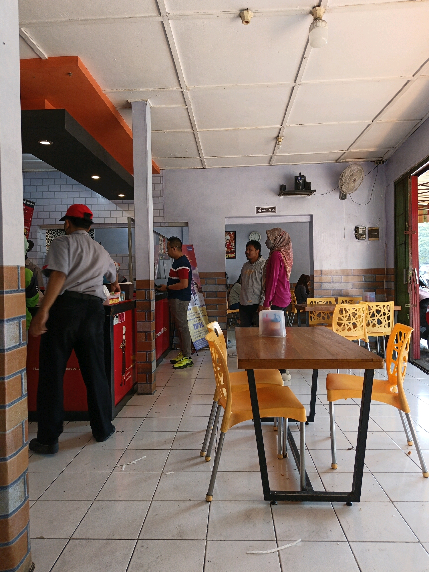 The image depicts the interior of a simple eatery, likely in Klaten, Indonesia, characterized by tiled floors, a suspended white ceiling with exposed beams, and bright orange and dark gray architectural elements. In the foreground, a wooden table with a metal frame is surrounded by bright yellow plastic chairs. To the left, a person wearing a red cap, gray shirt, and black pants is interacting with a red service counter. Further back, several other individuals are visible. Two men and a woman in a hijab are standing near a doorway. Another person is seated at a table in the background. A ceiling fan is mounted on the wall. The overall ambiance suggests a casual dining establishment.