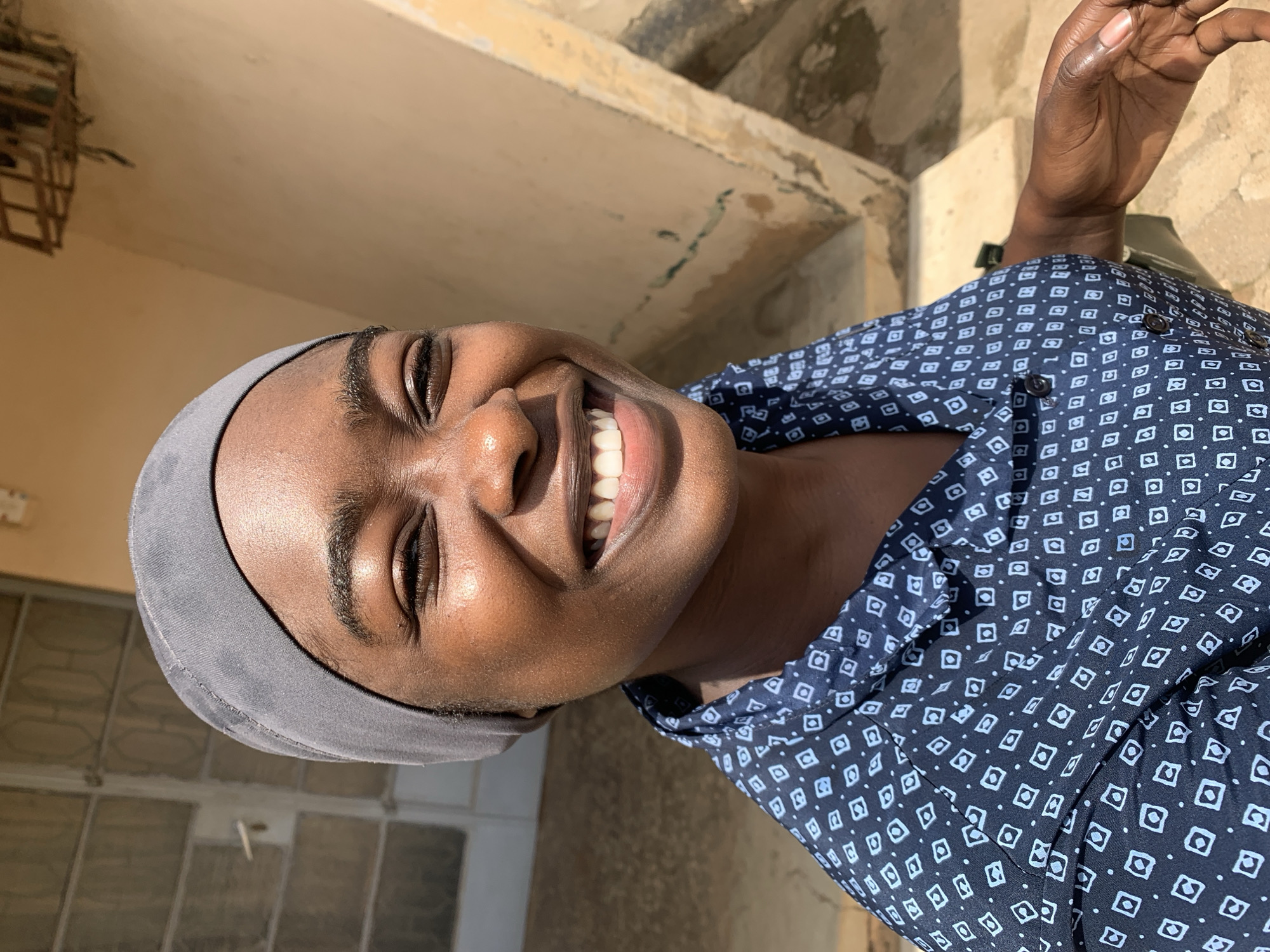 A medium shot photograph depicts a smiling woman with dark skin. She is wearing a dark blue collared shirt printed with a pattern of small white squares, and a grey headscarf. The background appears to be an exterior space with weathered walls and glimpses of an adjacent building. The location can be identified as Rafin Par, Nigeria. The lighting is bright, suggesting the photo was taken outdoors during the day. The woman's hand is partially visible in the upper right corner of the image.