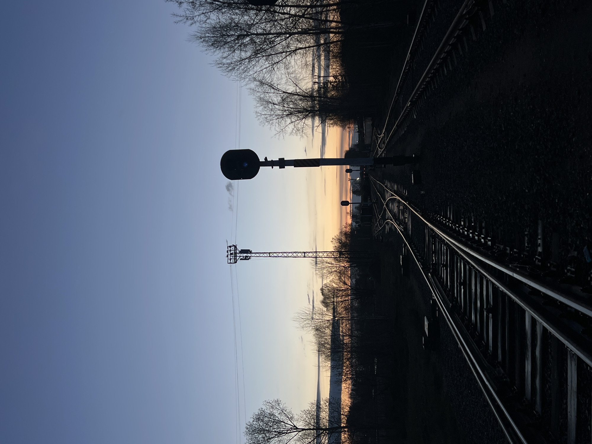 This media file, captured in Chaikovsky, Russia, depicts a serene outdoor scene at either sunrise or sunset, viewed along railroad tracks. The image is oriented vertically, with the sky dominating the left side, transitioning from a deep blue at the top to a warm gradient of light blue, pink, and orange near the horizontal center of the frame, indicating the horizon.
In the foreground and mid-ground, a network of dark railroad tracks stretches from the lower right, curving gently towards the horizon on the upper right side of the image. The tracks and the ground immediately surrounding them are heavily shadowed, appearing almost black, with reflections faintly visible on the rails.
A prominent railroad signal stands in the mid-ground, to the left of the tracks. It features a circular, dark lamp atop a pole with distinct yellow and black diagonal stripes. A faint wisp of vapor or smoke is visible near the lamp. Further to the left and slightly behind the signal, a tall, metallic lattice tower, likely a power or communication mast, stands silhouetted against the colorful sky.
Silhouetted bare trees line the horizon across the middle of the image, their branches stark against the fading light. The overall mood is quiet and atmospheric, characterized by the dramatic contrast between the illuminated sky and the dark, shadowed landscape elements. No people or active events are depicted, and no visible text can be discerned. The weather appears clear and calm.
