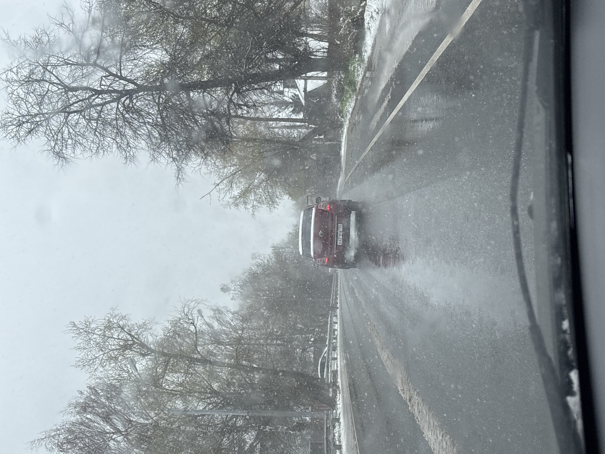 The image depicts a road scene from the perspective of a vehicle driving during a snowfall in Maksimkovo, Russia. The road surface is wet, with patches of slush or melted snow, and snowflakes are actively falling, creating a diffuse, wintry atmosphere.
Ahead, a dark red SUV, likely a Nissan, is visible, driving in the same direction. Its taillights are illuminated, and a slight spray of water or slush is visible behind its tires. The vehicle's rear license plate displays 'E471PP 750', with the '750' indicating Moscow Oblast, consistent with the specified location.
Bare, deciduous trees, some with a light dusting of snow on their branches, line both sides of the road, suggesting winter or very early spring. A silver metal guardrail runs along the right side of the road. The sky is overcast and bright, indicating daytime conditions. The foreground on the right shows a blurred portion of the vehicle's interior, such as a window frame or dashboard, from which the photo was taken. The scene conveys the conditions of driving through a snowy landscape.