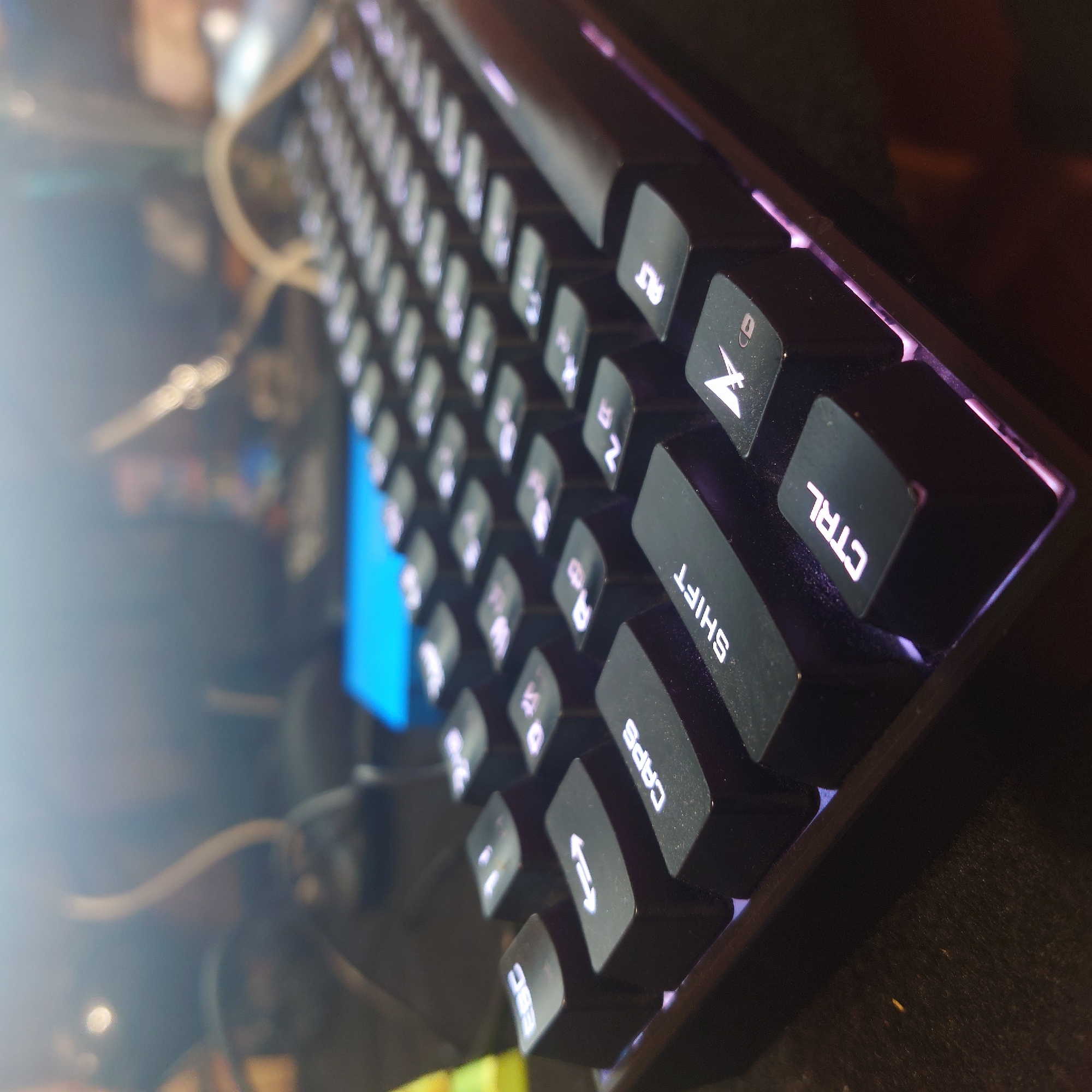 The image shows a close-up, angled view of a black mechanical keyboard with backlit keys, set against a blurred background. The keys visible include ESC, CAPS, SHIFT, CTRL, and several number and symbol keys. The lighting suggests a dimly lit environment, with purple backlighting emanating from the keyboard. The overall impression is of a gaming or computer setup. There is no discernible information in the image that would indicate its location in Belorechensk, Russia, or any specific environmental context beyond the immediate desk area.