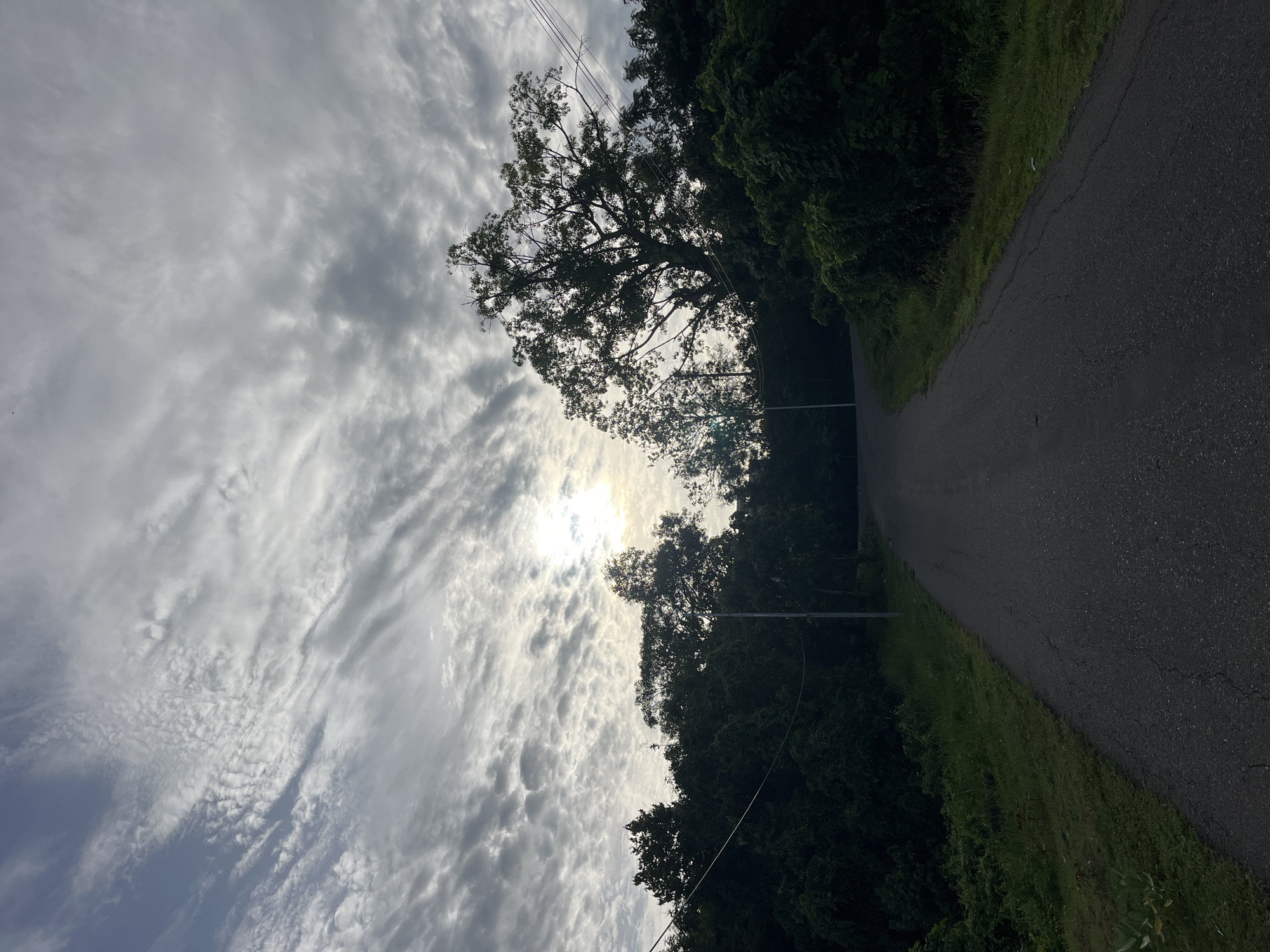 The image shows a road in a rural or semi-rural location. The road appears to be asphalt, with green vegetation along both sides. The sky is heavily clouded, with the sun visible through the clouds, creating a bright glare. There are trees lining the road, silhouetted against the sky. Power lines are also visible, running parallel to the road. The location is stated to be Lhokseumawe, Indonesia, but there are no specific features in the image that confirm this information without additional context. There are no people visible in the image.