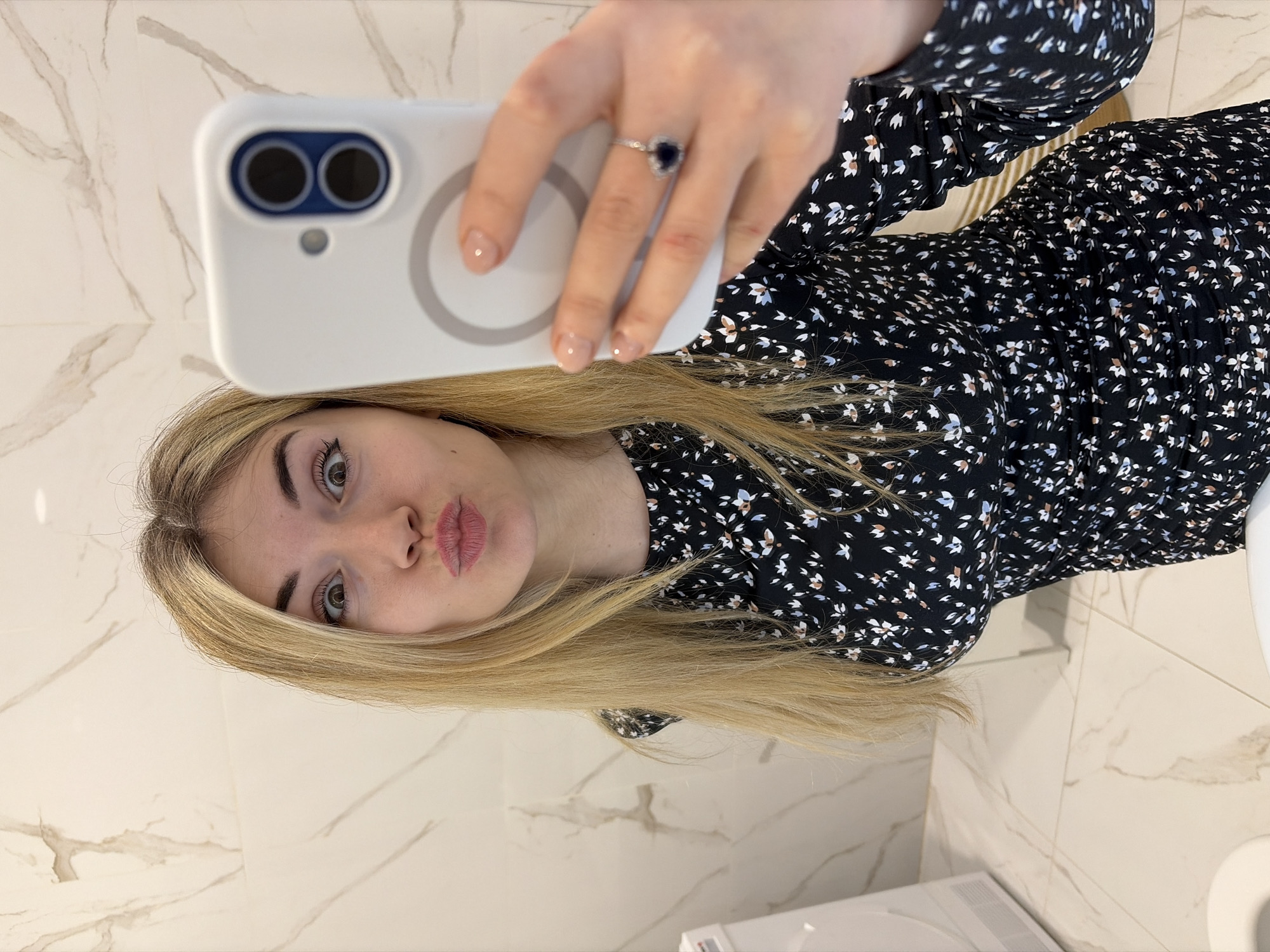 A blonde female subject is depicted taking a self-portrait photograph (selfie) in what appears to be a residential bathroom in Budva, Montenegro. The subject is holding a white smartphone, likely an iPhone model with a dual-camera system, encased in a white protective cover featuring a circular MagSafe ring, in her right hand. A ring with a dark, heart-shaped stone is visible on her left ring finger. She is wearing a black long-sleeved dress with a repeating pattern of small white, blue, and brown floral elements. Her facial expression includes wide-open eyes and pursed lips. The background consists of large format white tiles with subtle grey-brown veining, covering both the walls and floor, indicative of a modern bathroom. A white appliance, possibly a washing machine or dryer, is partially visible on the left side of the frame, and the rim of a white toilet bowl is visible in the lower left foreground.