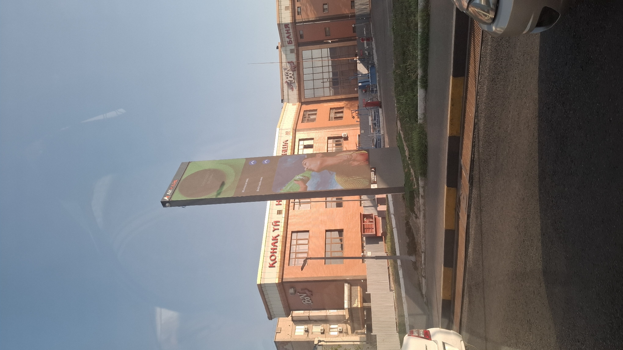 The image shows a street scene in the city of Shymkent, Kazakhstan. A tall, vertical digital billboard is prominently displayed in the foreground, showcasing an advertisement with an image of a woman drinking from a bottle and text in Russian, including "AKFA" at the top. The billboard is positioned in front of a modern, multi-story brick building with large windows and signage that reads "КОНАК ҮЙ" and "BN" on one section, and "БАЊЯ" on another. The building appears to be a commercial or entertainment complex. A street with a yellow and black curb runs in front of the building. A white car is partially visible on the left side of the image, and the front of a silver car is visible on the right. The sky is clear and blue, suggesting it is daytime. The overall environment is urban.