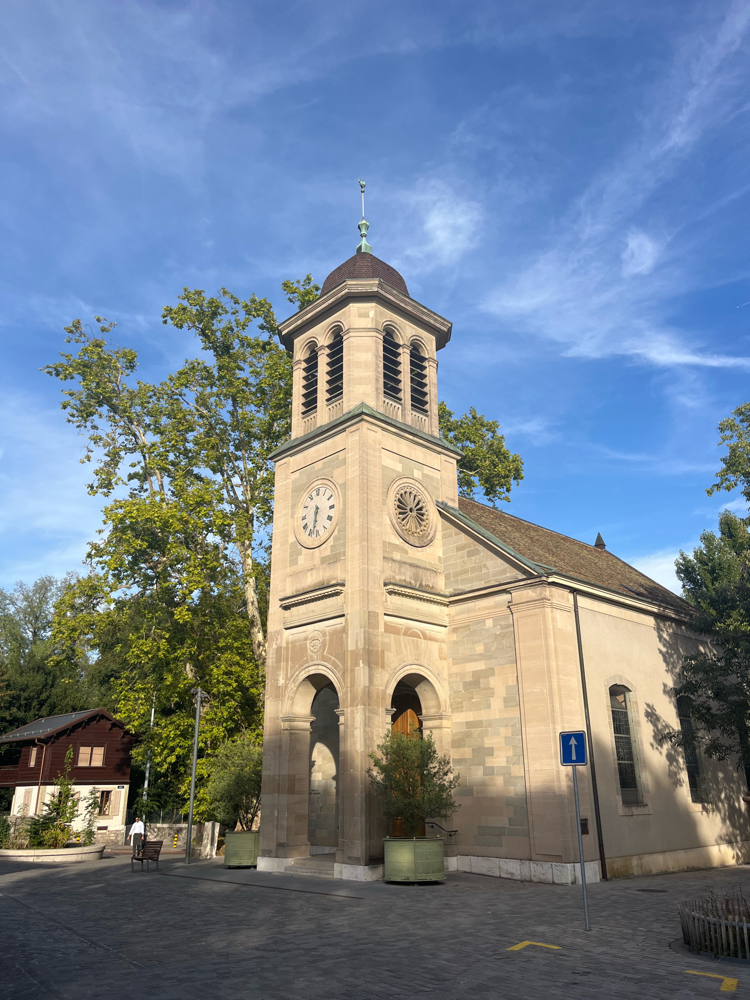 This is a full shot of a church with a bell tower, located in the city of Geneva, Switzerland. The church is constructed of light-colored stone blocks. The bell tower has a clock face with Roman numerals and a decorative circular window above an arched entrance. A green-domed roof caps the tower. The main body of the church has a sloping, tile roof.
To the left of the church, a two-story wooden house with a dark brown exterior and light-colored trim is visible. A single person wearing a white shirt and dark pants is walking on a paved cobblestone area in front of the house and the church. A park bench is placed in the foreground near the person. There are trees with green foliage surrounding the building and a blue directional sign with a white arrow pointing upwards. The sky is clear blue with scattered wispy white clouds.
