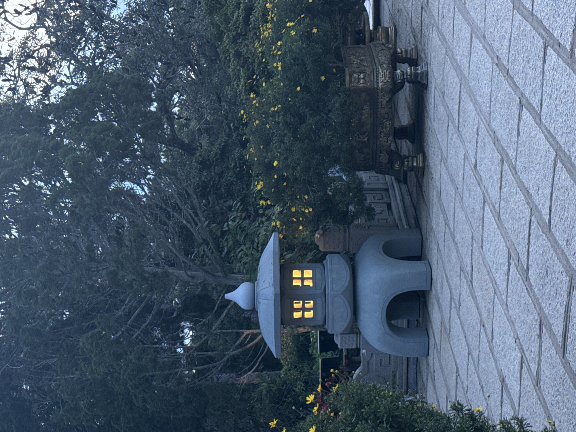 This image captures an outdoor garden or temple courtyard, identified as being in Da Lat, Vietnam, under low ambient light conditions consistent with twilight. A central, grey, multi-tiered stone lantern features a pointed finial and four rectangular openings on its body, from which warm yellow light emanates. The lantern's base includes four arch-like cutouts. To the right of the lantern, a large, dark green leafy plant, profusely dotted with small bright yellow flowers, rests within an ornate, dark bronze-colored metal planter detailed with relief carvings and supported by four intricate legs. A similar flowering plant is partially visible to the left. The ground is paved with rectangular stone blocks laid in a grid pattern. The background is dominated by dense foliage of various trees, some displaying dark green leaves and others exhibiting a more skeletal branch structure. Indistinct stone architectural elements are visible behind the lantern and the large planter. No human figures are present.