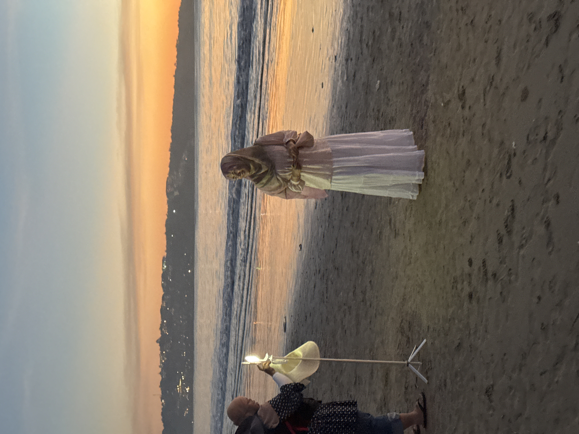 The image shows a scene on a beach at sunset. A statue of a veiled figure is positioned on the sand, facing the water. A person is standing on the beach using lighting equipment. A stick with a tripod base is positioned near the subject. The sky is illuminated by orange and yellow hues. The photograph location is on a beach in Kuta Selatan, Indonesia.