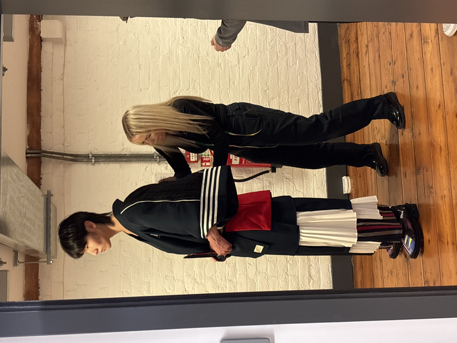The scene depicts an indoor fitting or styling session within a studio or creative workspace in the city of London, United Kingdom. A person with dark, medium-length hair stands in profile, facing left, wearing a complex, multi-layered avant-garde outfit. This ensemble features a dark kimono-style jacket with white stripes on the lower left sleeve and a textured dark panel on the upper back, a visible red fabric element beneath, a white pleated skirt-like garment, and vertically striped trousers in black, red, and white. They wear chunky black sneakers with colorful accents and a prominent Puma logo. To their right, a blonde-haired woman, dressed in a black long-sleeved top and dark trousers with a yellow drawstring detail, stands facing the first individual, actively adjusting the back of their jacket. A third person's right arm and shoulder, wearing a grey jacket, are partially visible on the far right. The background consists of a white-painted exposed brick wall, a polished wooden floor, and exposed metal conduit on the ceiling. A red fire extinguisher is mounted on the brick wall.