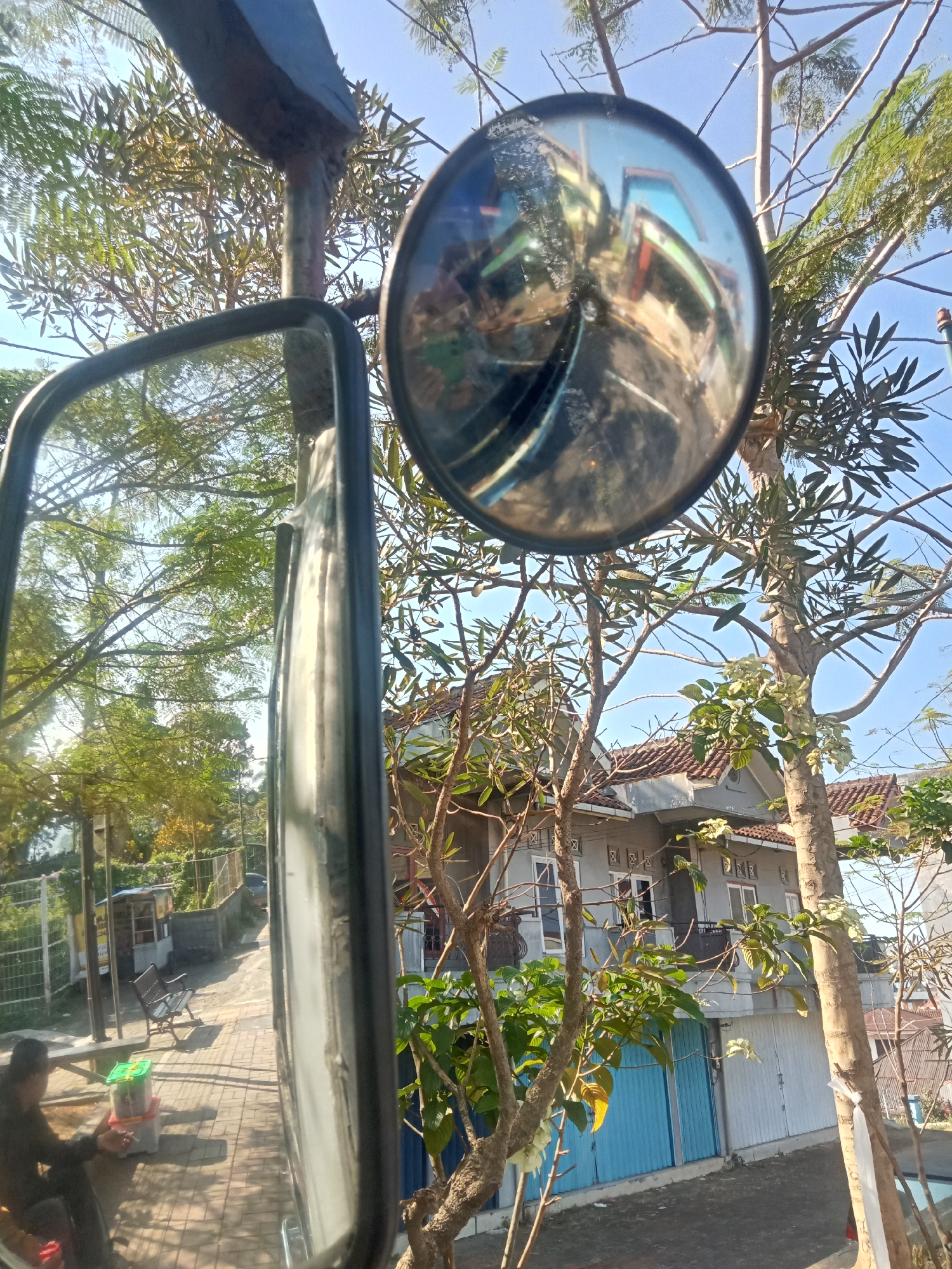 A wide-angle shot captures a street scene in Jaraksari, Indonesia, as reflected in two mirrors. The primary mirror, positioned on the left, shows a paved sidewalk with brick patterns. A person in dark clothing sits on a bench, holding a cigarette and a green container. Behind them, a small kiosk and more benches are visible. The secondary, round mirror on the right reflects a street with colorful vehicles and a clear blue sky. Trees with green foliage surround the scene, with a light gray house featuring a red-tiled roof and blue and white garage doors in the background. The sunlight casts shadows, indicating it is daytime.