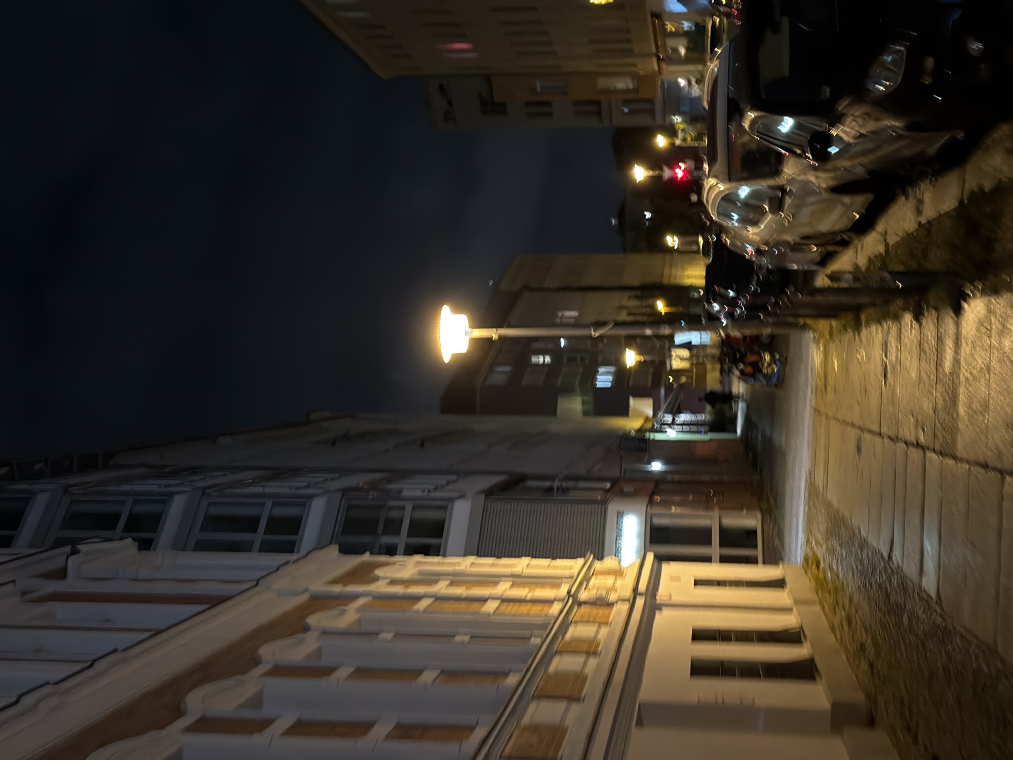 The image depicts a street scene in Berlin, Germany at night. The perspective is from a sidewalk looking down a street lined with buildings and parked cars. The buildings on the left are illuminated by streetlights, revealing ornate architectural details. On the right, parked cars are visible, their shapes softened by the low light and motion blur. Several streetlights are also present, emitting a warm glow. The sky is dark and cloudy, suggesting nighttime conditions. The street appears to be paved with cobblestones or similar material, and there is a sidewalk with visible tiles. The overall atmosphere is that of a quiet urban street after dark.